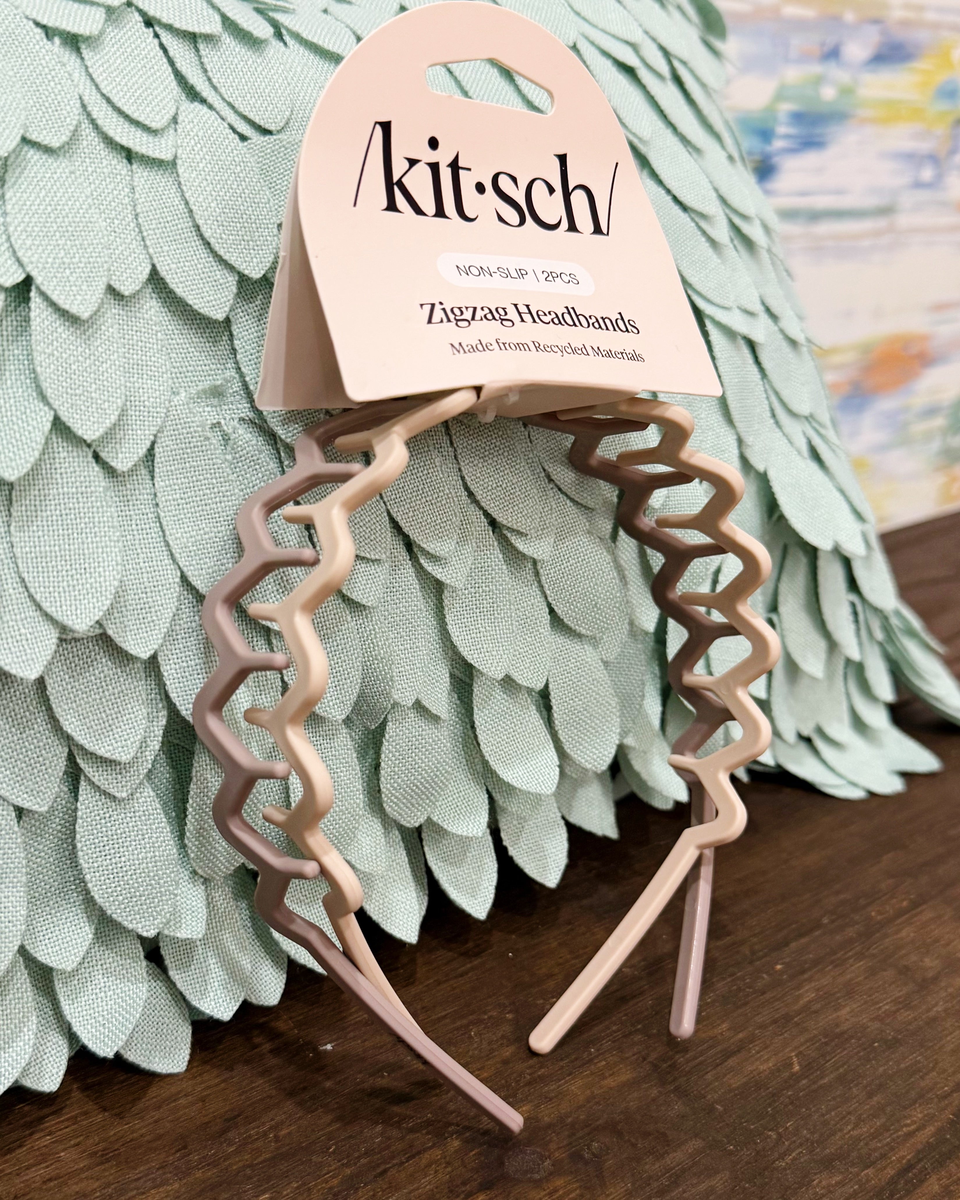 A set of three Kit'sch zigzag headbands in dusty pink, beige, and taupe, attached to a paper hanger card and displayed on a wooden surface in front of a mint green feather pillow.