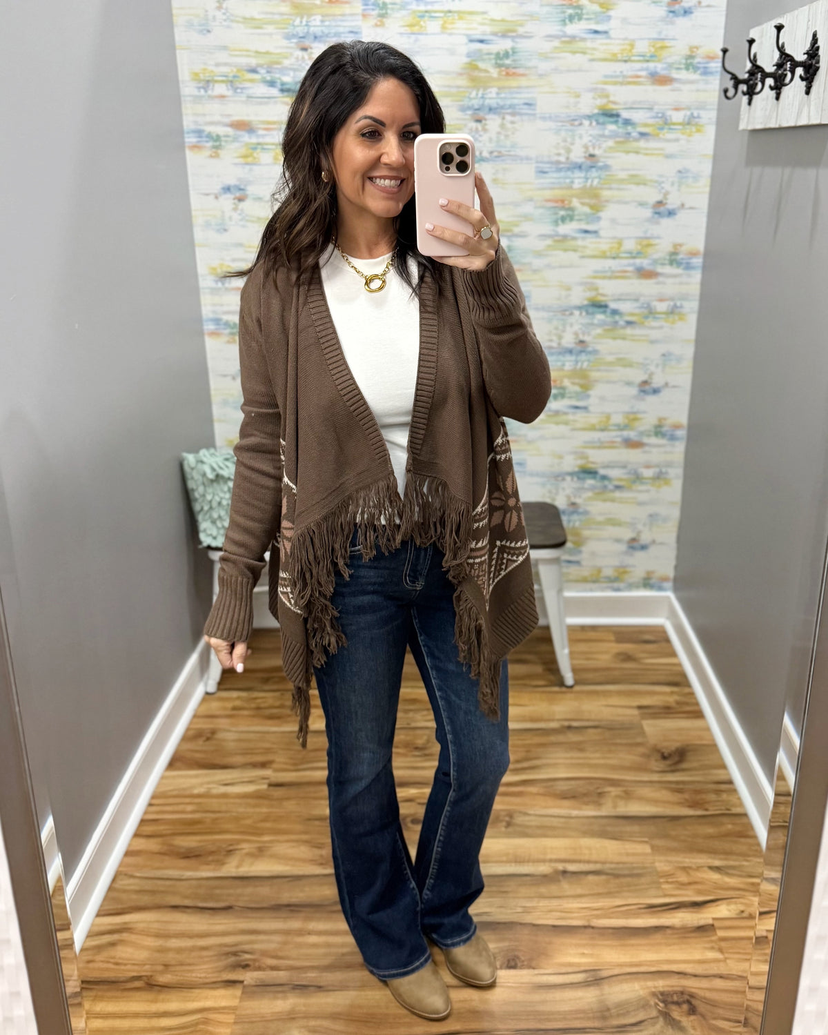 A woman takes a mirror selfie in a fitting room wearing a brown open-front fringed cardigan over a white top, blue jeans, and tan boots.