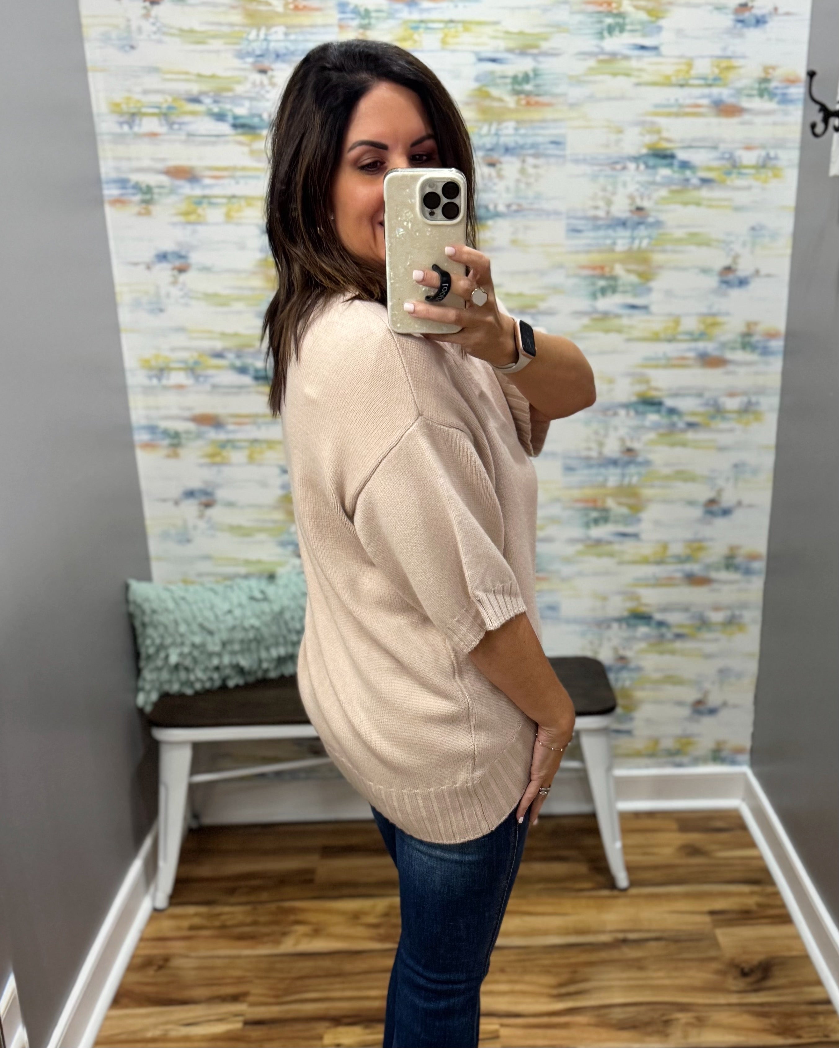 A woman takes a mirror selfie in a fitting room wearing an oversized light beige knit sweater and dark blue jeans.