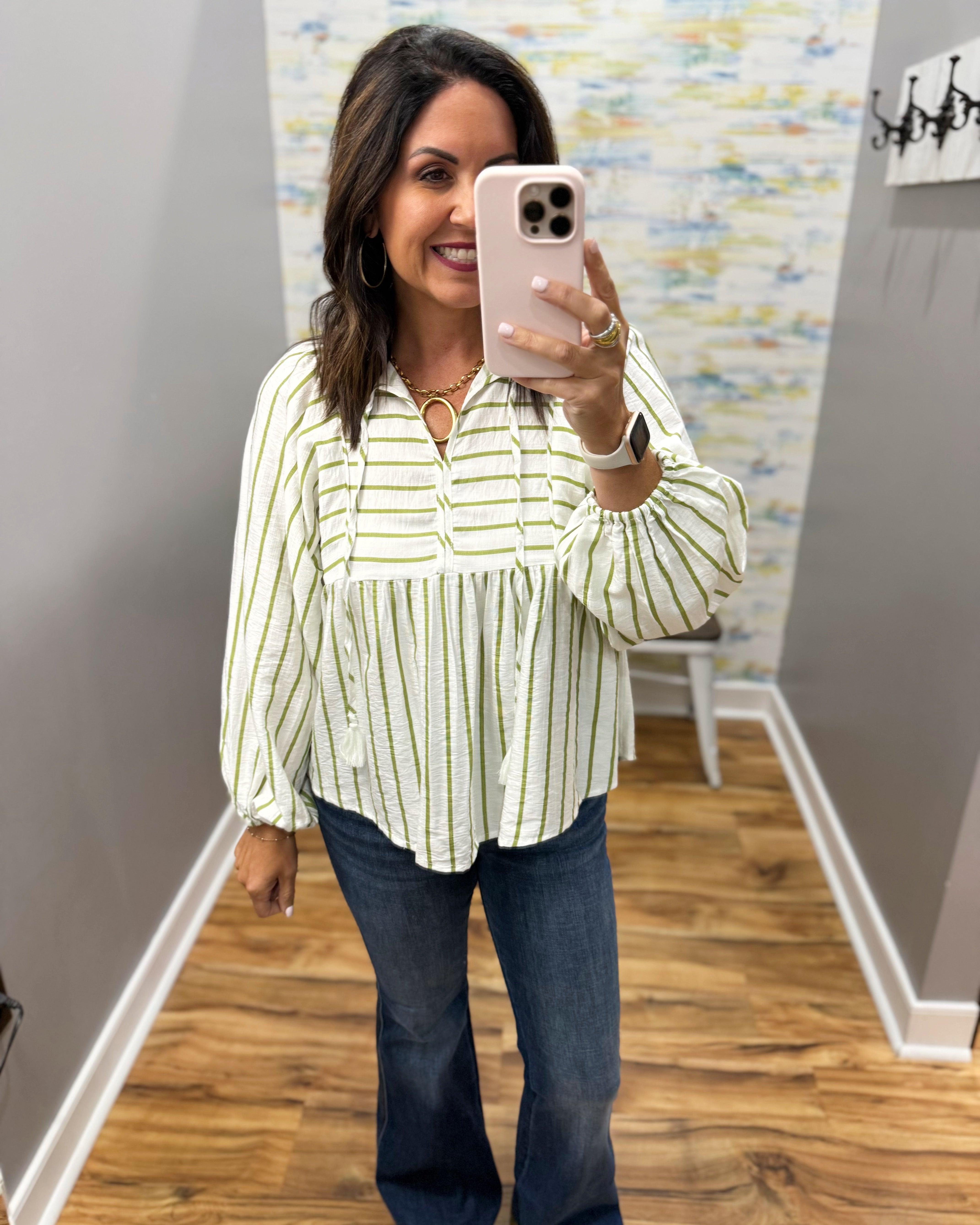 A woman takes a mirror selfie wearing a cream-colored blouse with thin green vertical stripes and dark blue jeans in an indoor hallway.