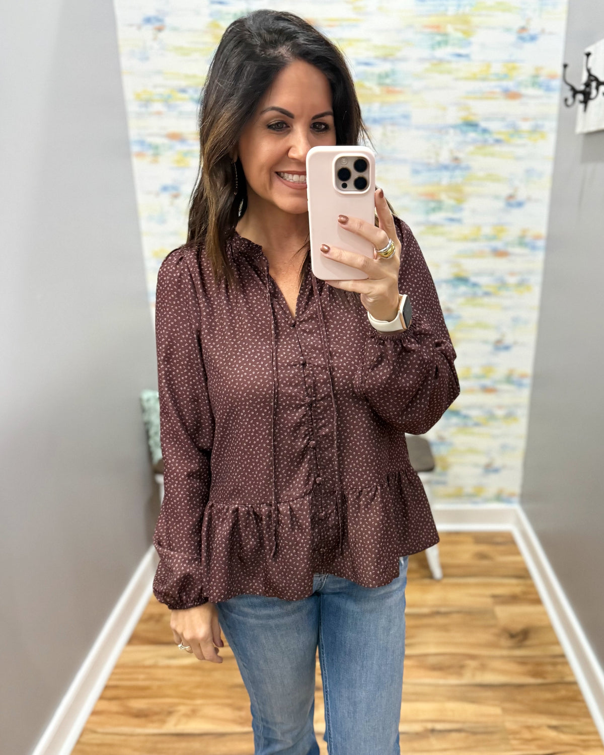 A woman takes a mirror selfie wearing a long-sleeve, button-front blouse with a ruffled front and peplum hem in a dark brown color with a small white floral pattern, paired with blue jeans.
