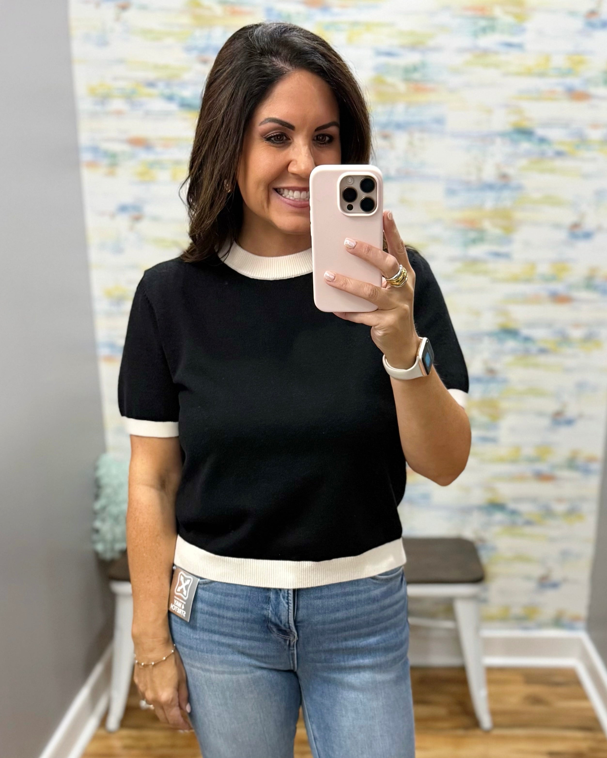 A woman takes a mirror selfie wearing a black short-sleeve knit top with white ribbed trim at the crew neck, sleeve cuffs, and hem, paired with blue jeans.