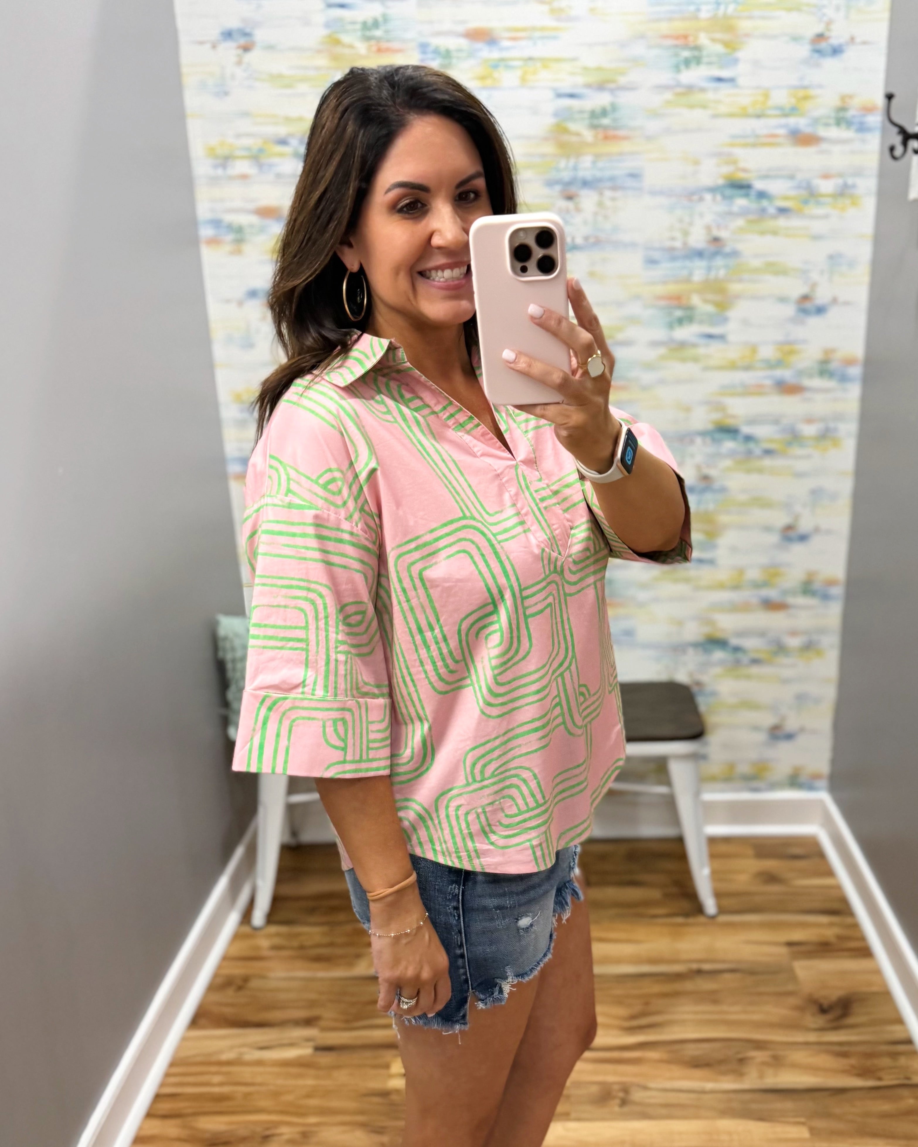 Woman taking a mirror selfie wearing a pink and green patterned shirt and denim shorts.
