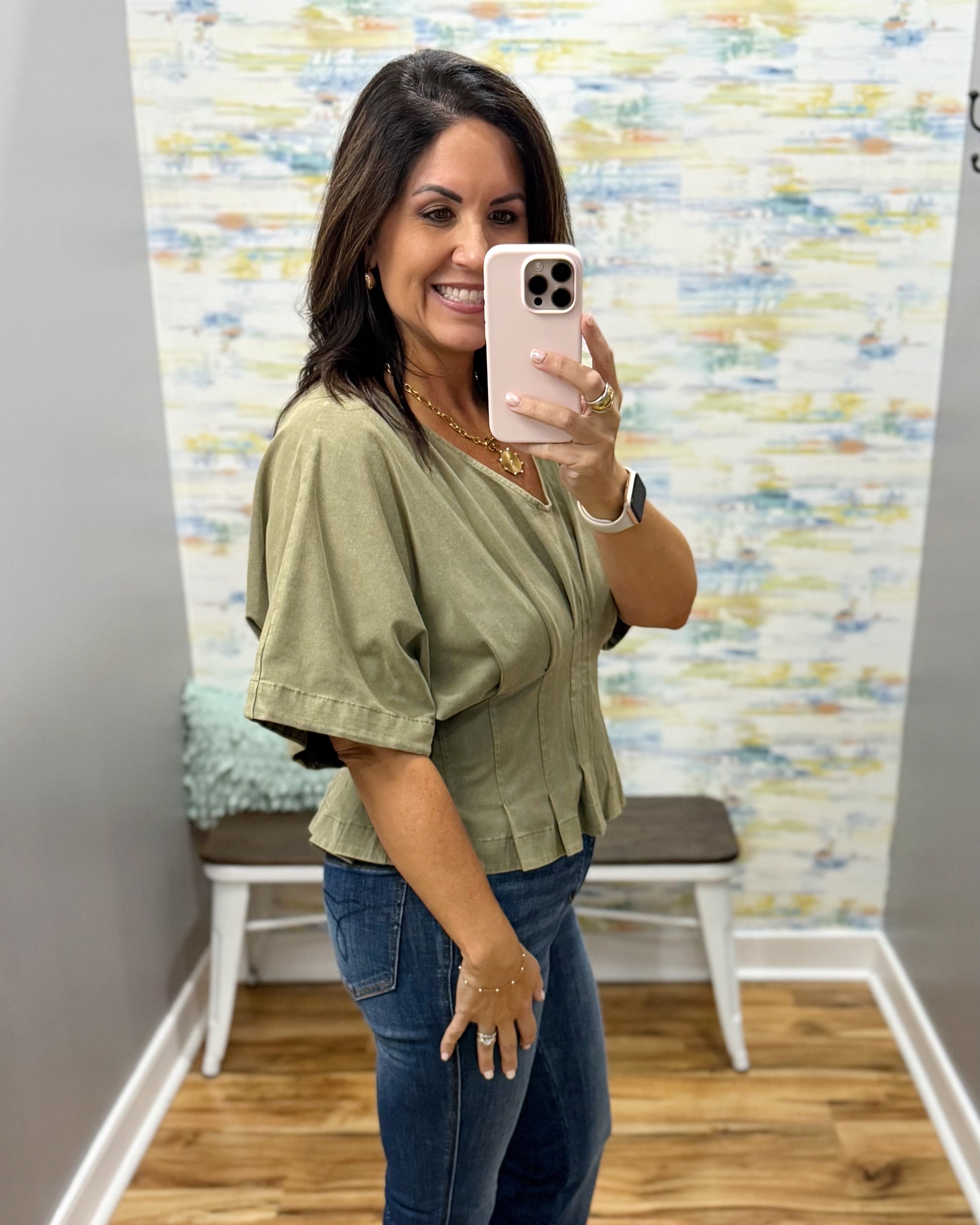 A woman takes a mirror selfie wearing an olive green short-sleeve blouse with pleated detailing and blue jeans in a fitting room with a patterned wall and wooden floor.