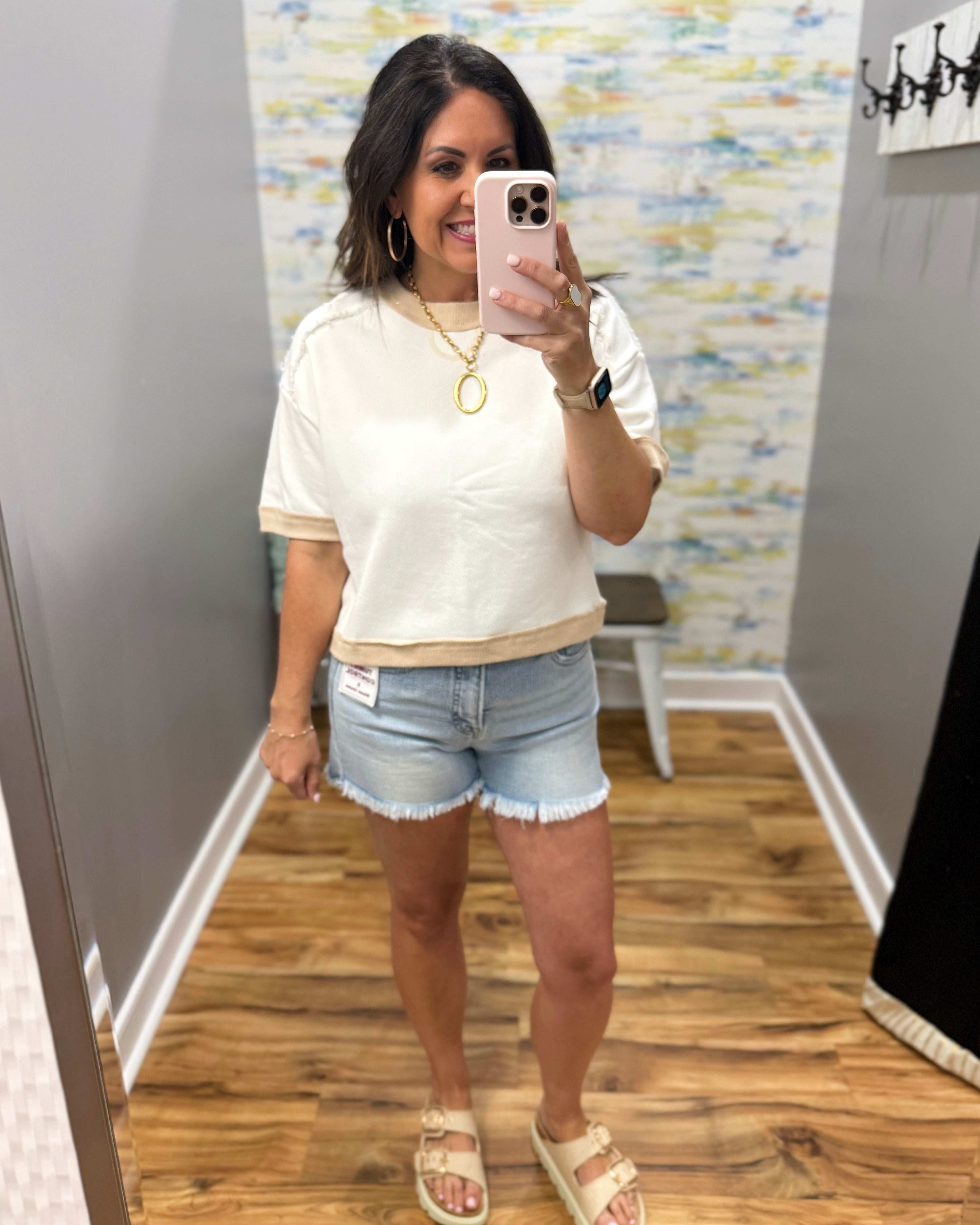Woman taking a mirror selfie wearing a white top and denim shorts in a room with a colorful wall.