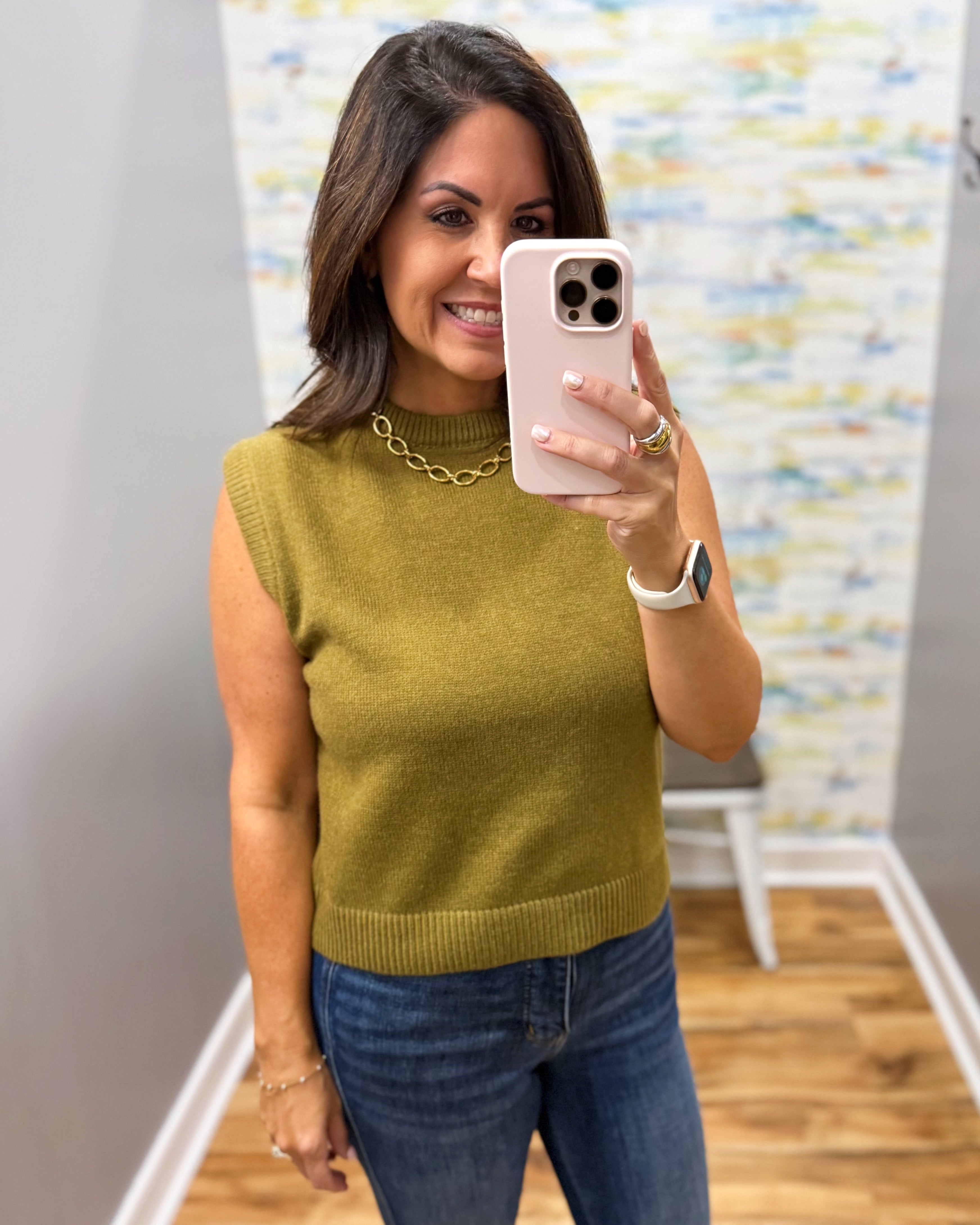 A woman taking a mirror selfie wearing a sleeveless olive green knit sweater vest with blue jeans and a gold chain necklace.