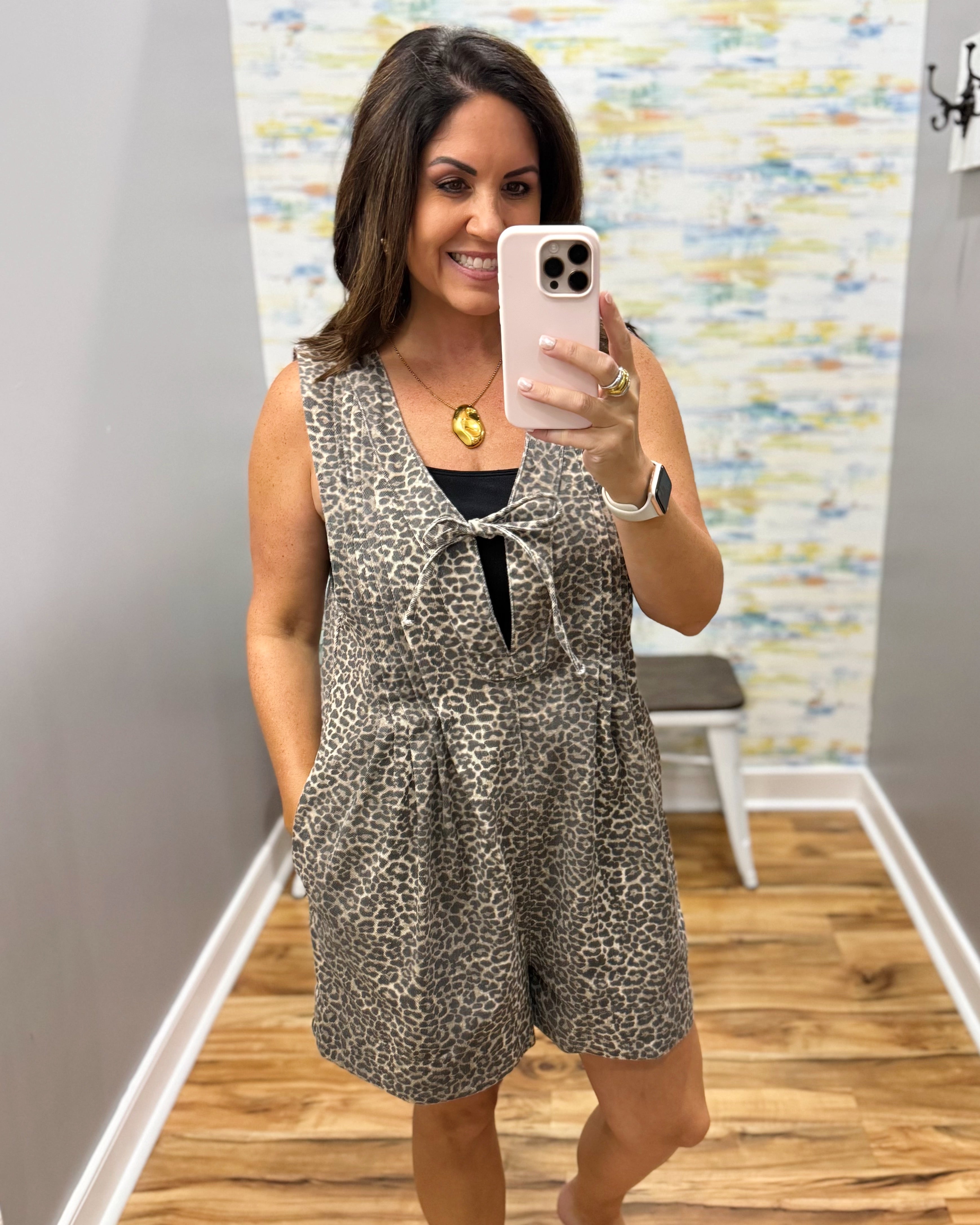 A woman takes a mirror selfie wearing a leopard-print sleeveless romper with a tie neckline over a black top. The setting is a home interior with wood floors and a patterned wall.