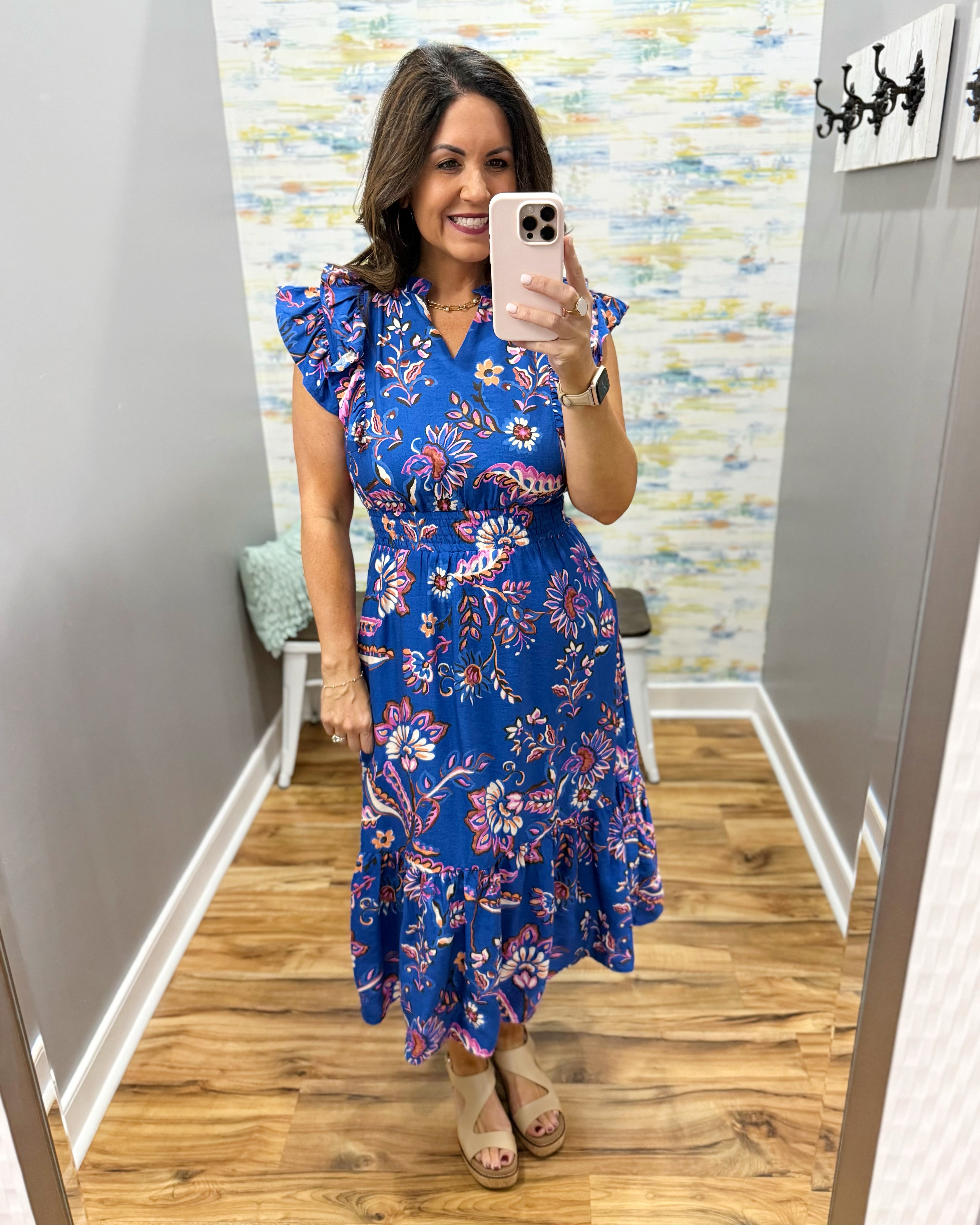 Woman taking a mirror selfie wearing a blue floral dress in a room with a patterned wall.