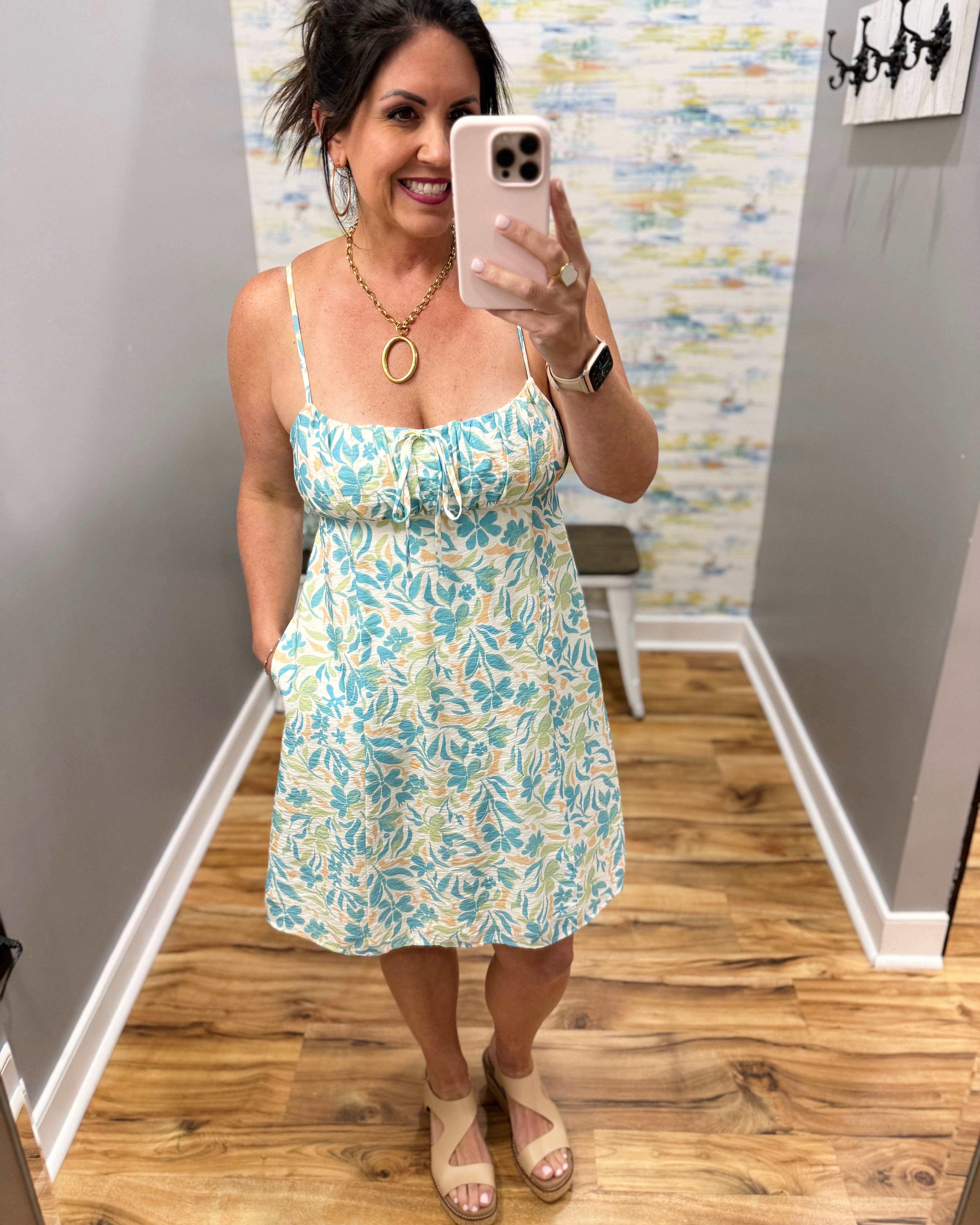 Woman taking a mirror selfie wearing a floral dress in a room with wooden flooring and a colorful wall.