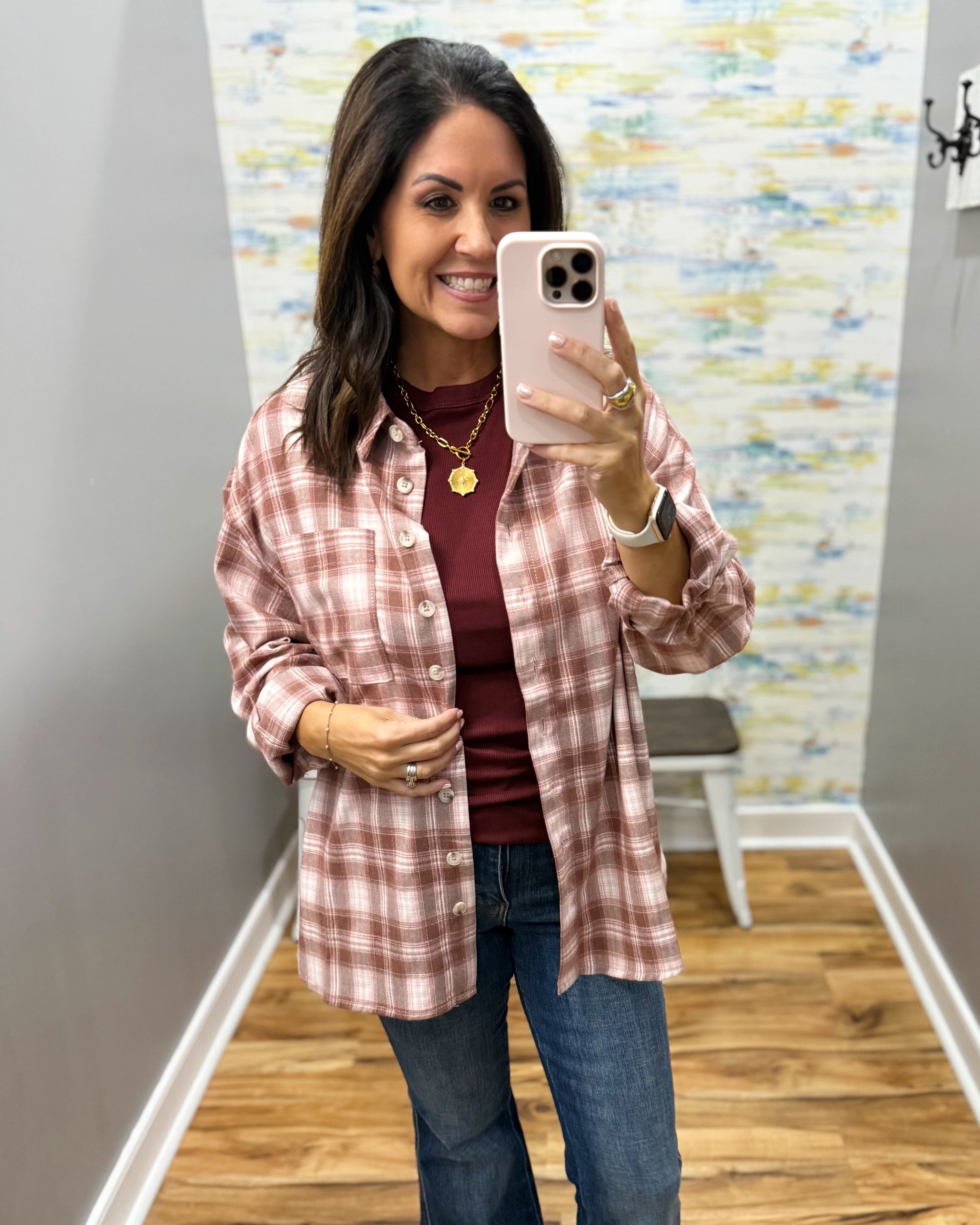 A woman takes a mirror selfie wearing a dusty rose and white plaid button-up shirt over a dark red top, paired with blue jeans. She is standing in a hallway with a patterned accent wall and wood floors.