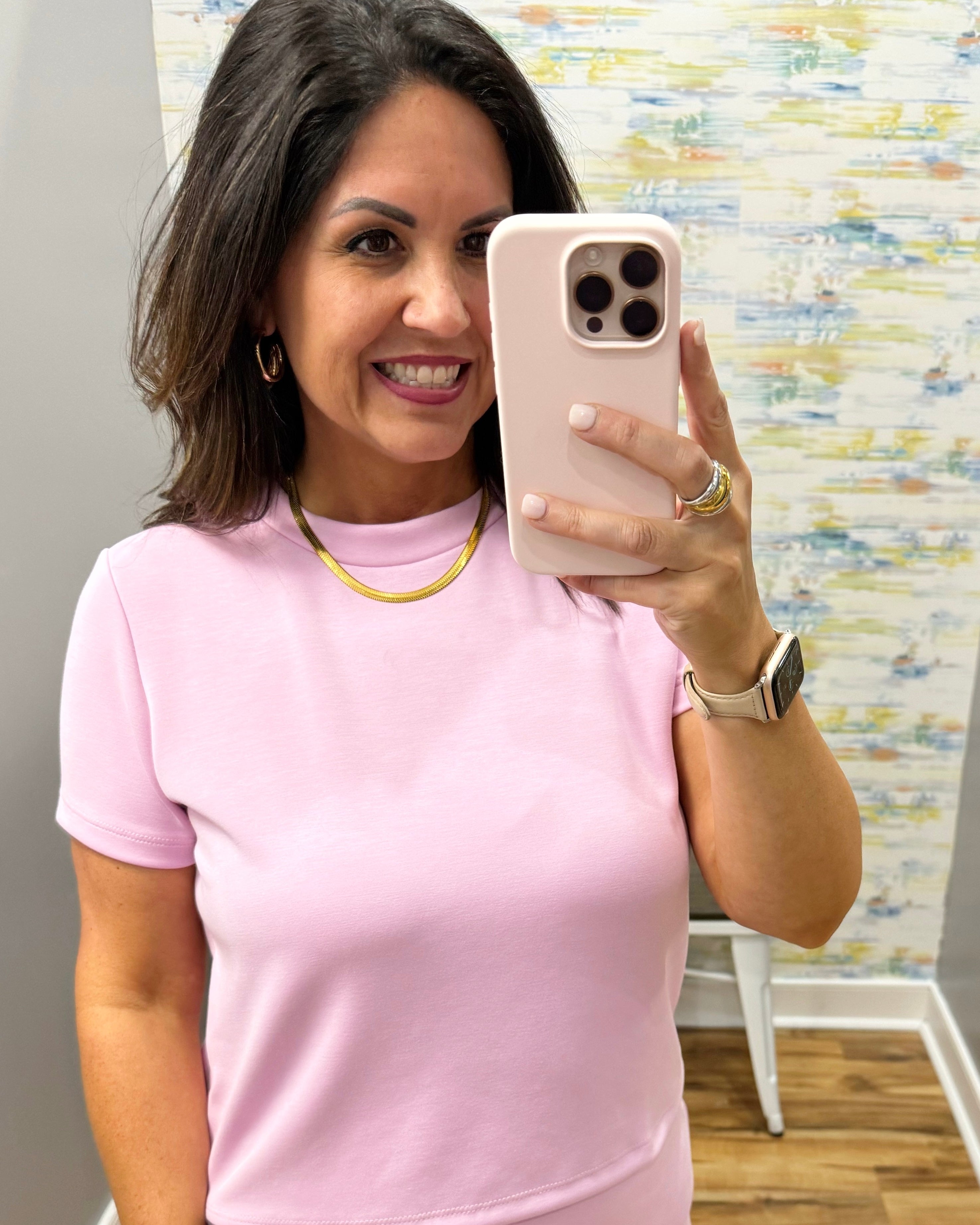 Woman taking a mirror selfie wearing a pink top with a colorful wall in the background.