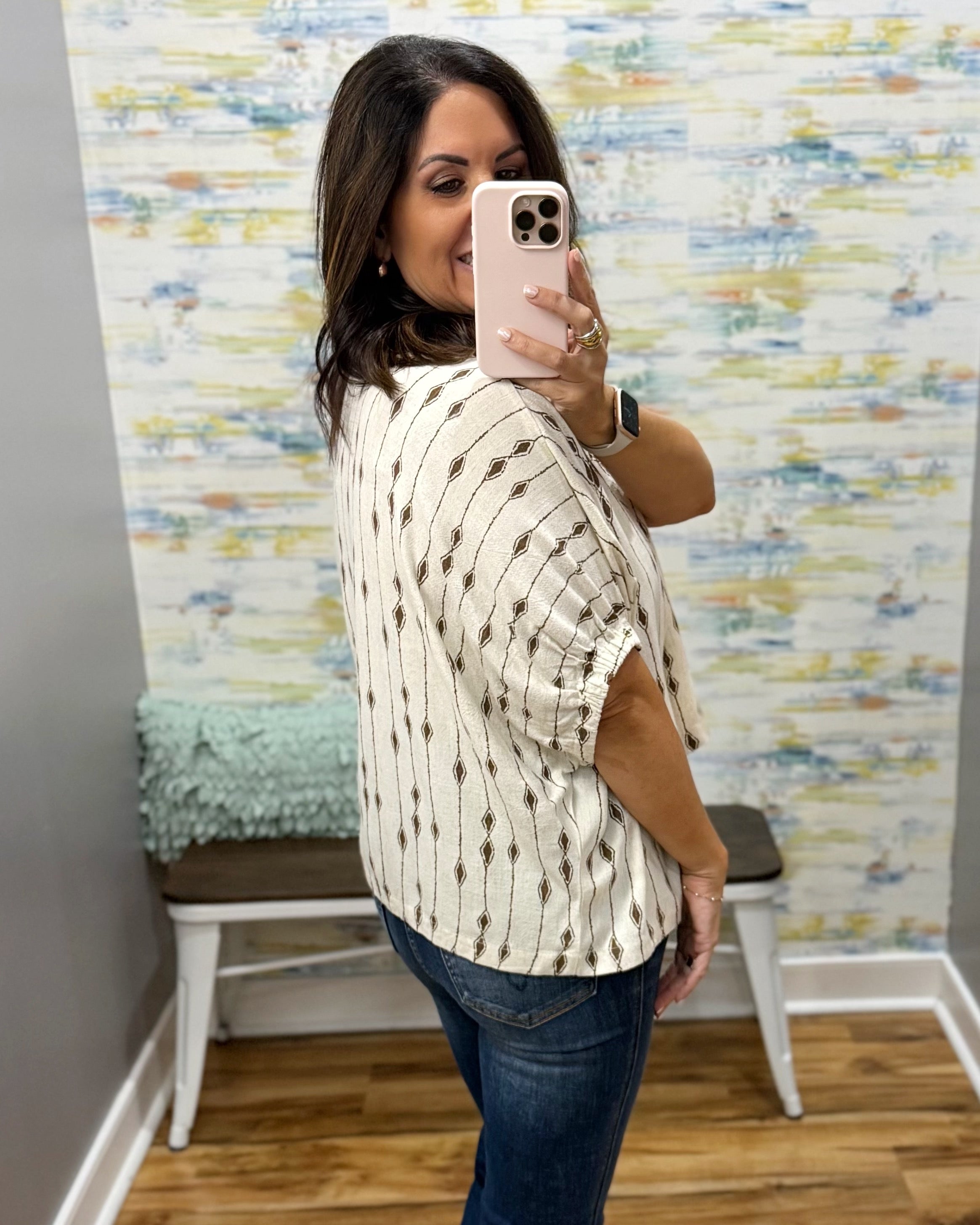 A woman takes a mirror selfie in a fitting room wearing a cream-colored short-sleeve top with a brown geometric pattern and blue jeans.