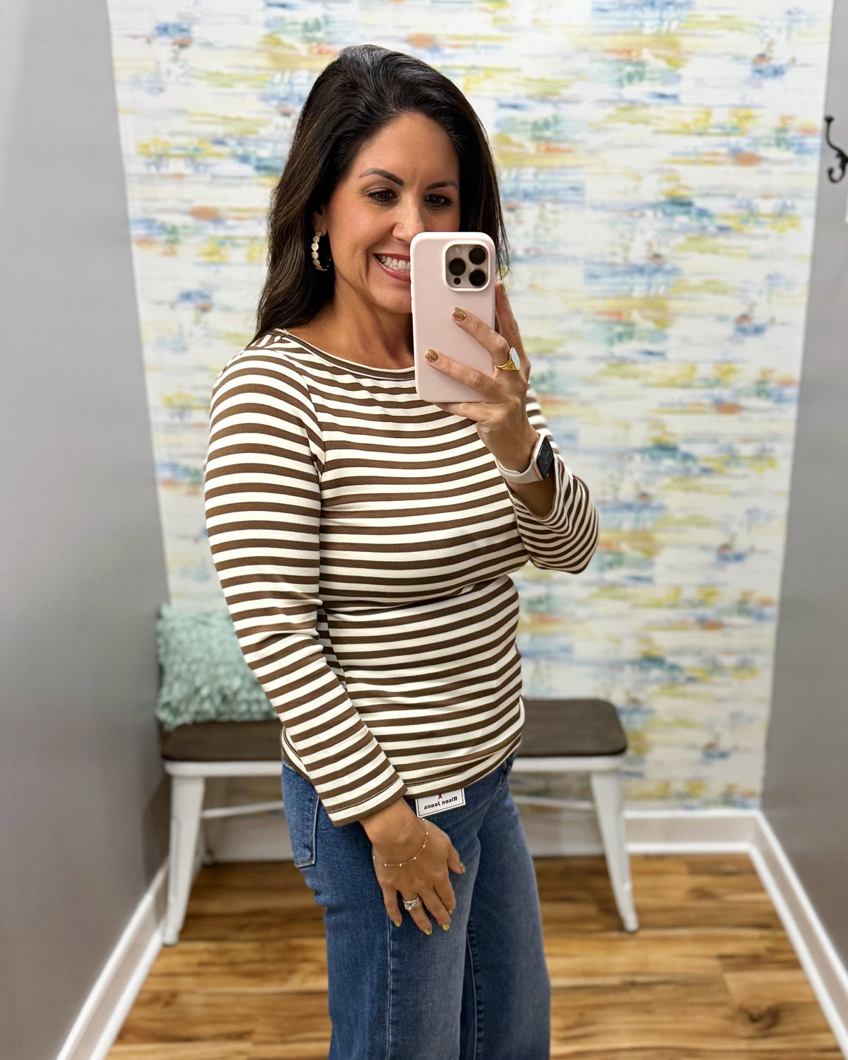 A woman takes a mirror selfie wearing a brown and white horizontally striped long-sleeve top and blue jeans in a fitting room with a patterned wallpaper background.