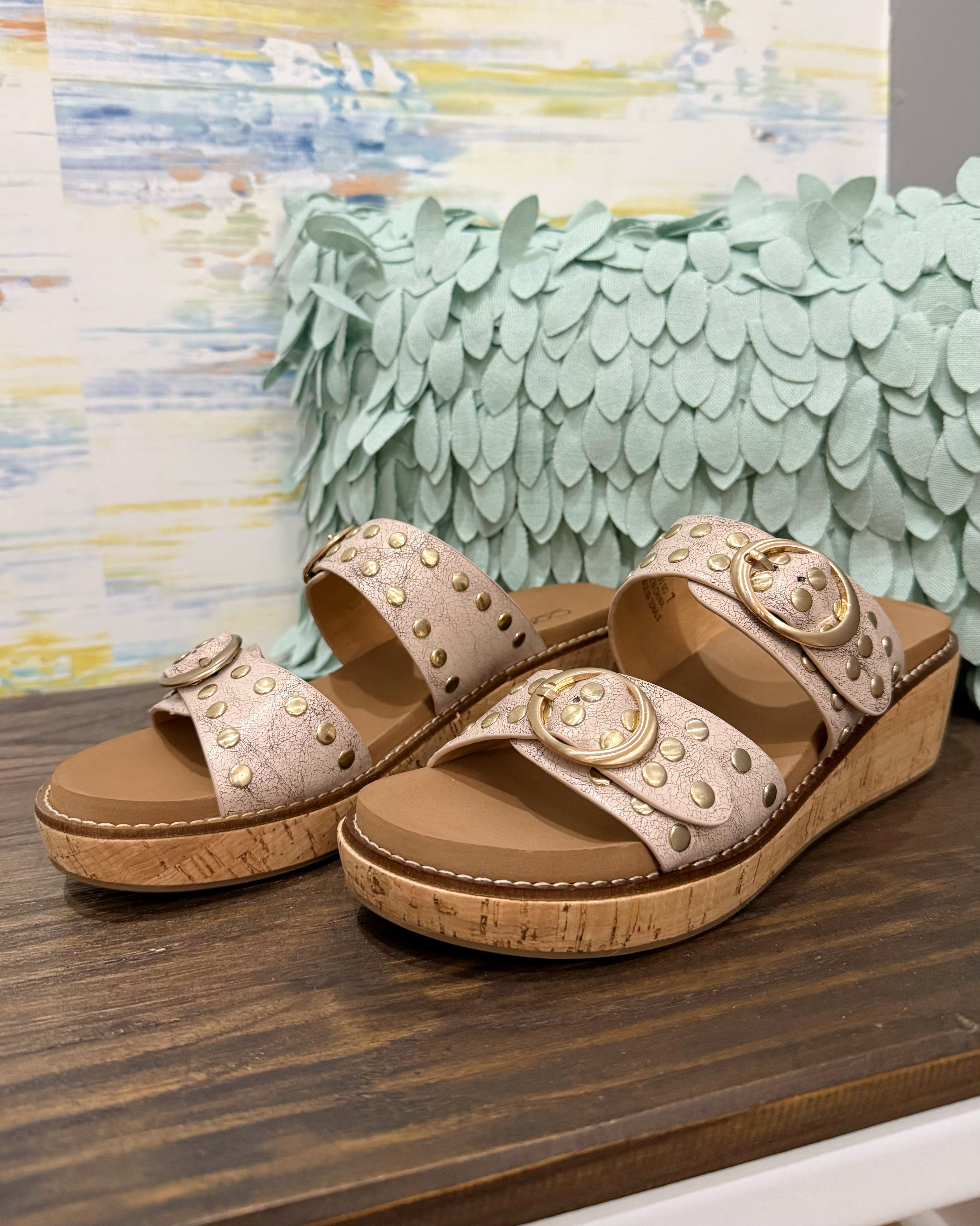 Pair of beige sandals with gold accents on a wooden surface with a decorative background.