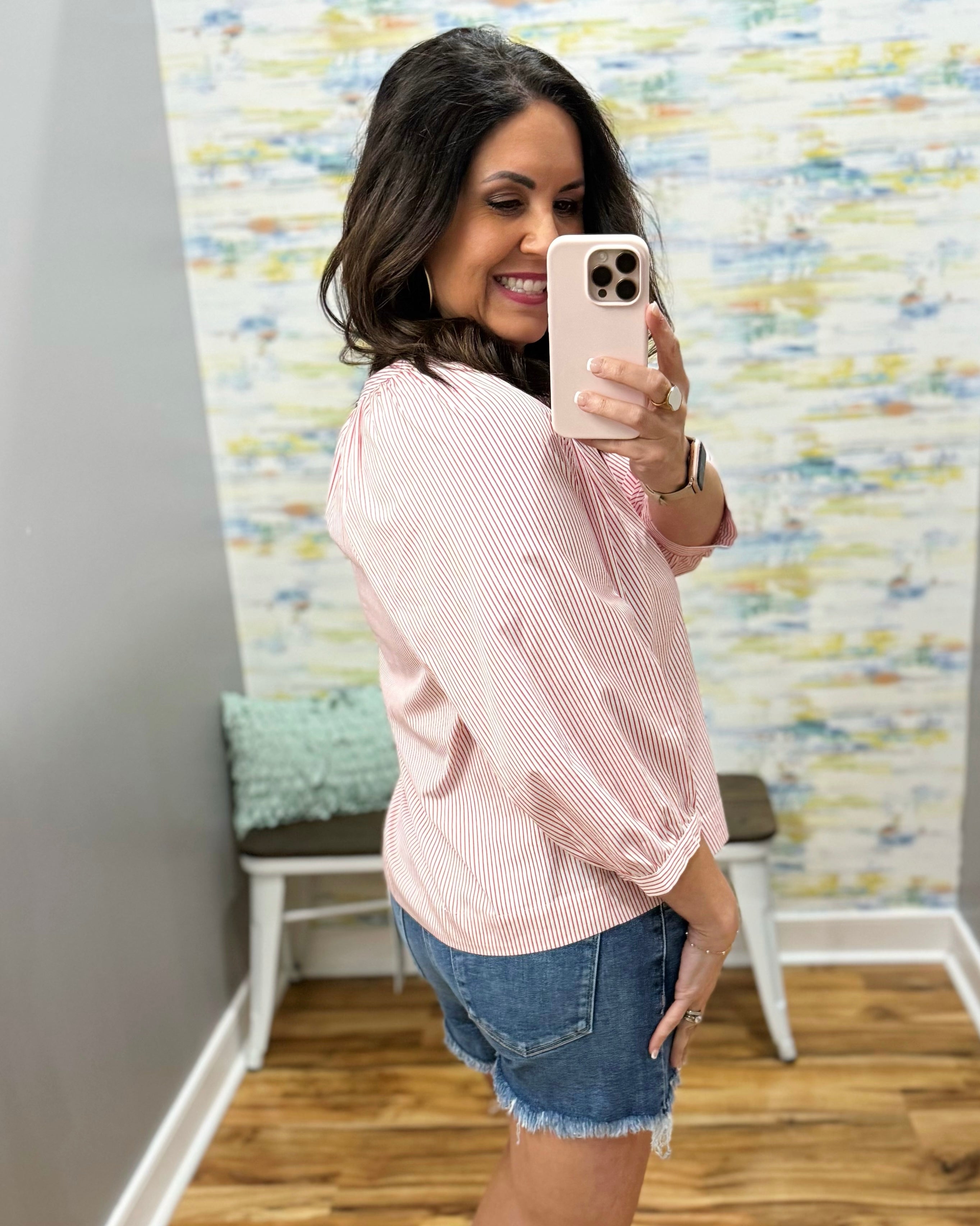 Woman taking a mirror selfie wearing a pink striped shirt and denim shorts in a room with a colorful textured wall.