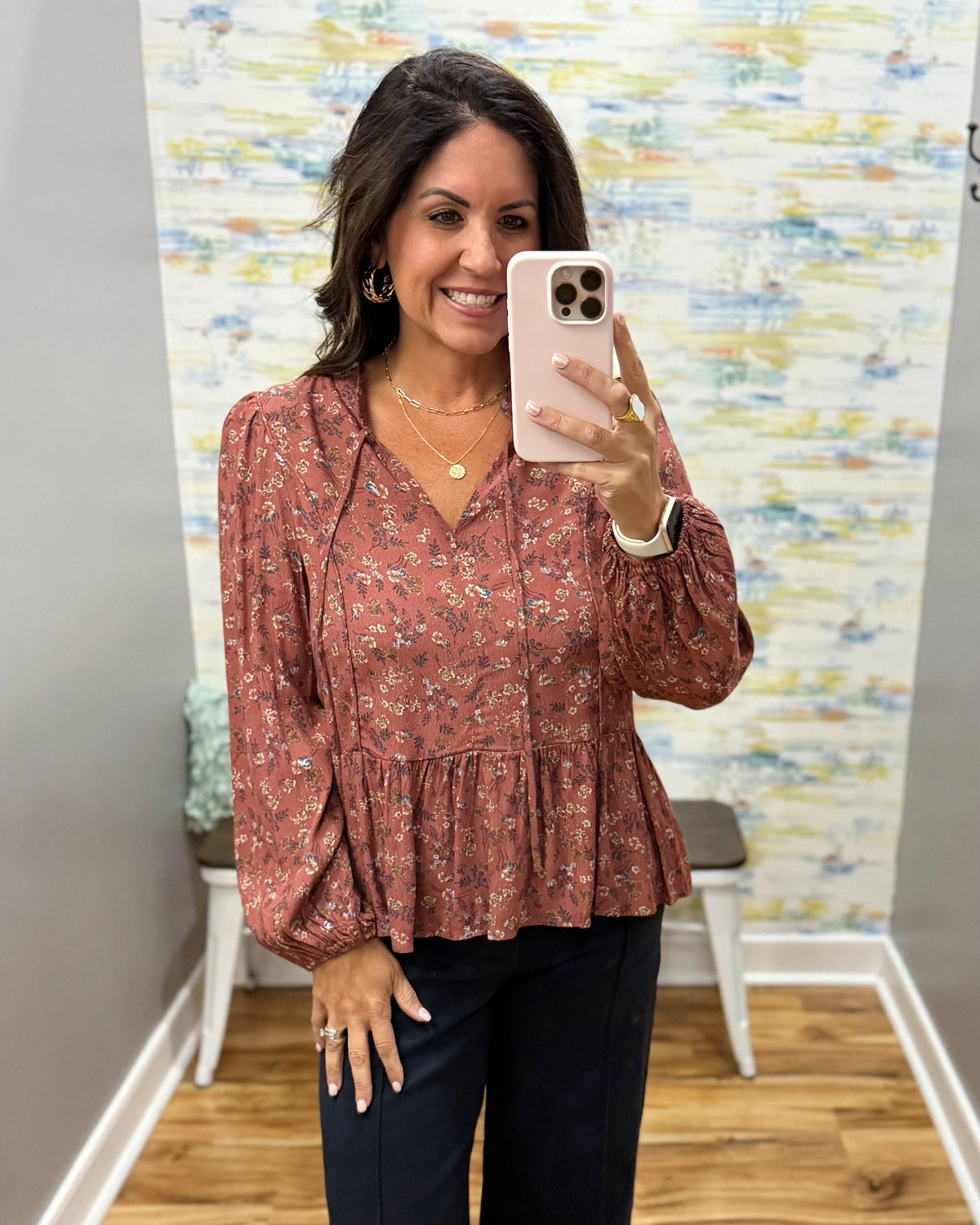 A woman takes a mirror selfie wearing a rust-colored long-sleeve blouse with a small floral print and dark pants in a fitting room.