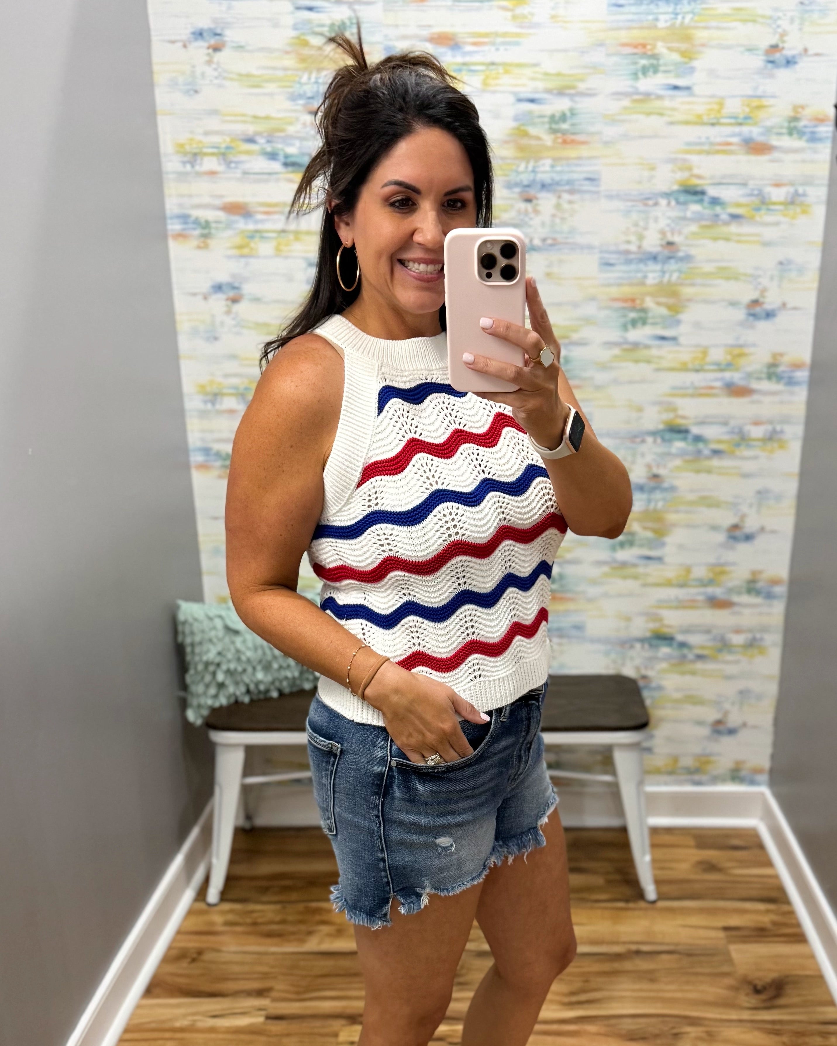 Woman taking a mirror selfie wearing a sleeveless top with red and blue wavy patterns and denim shorts.