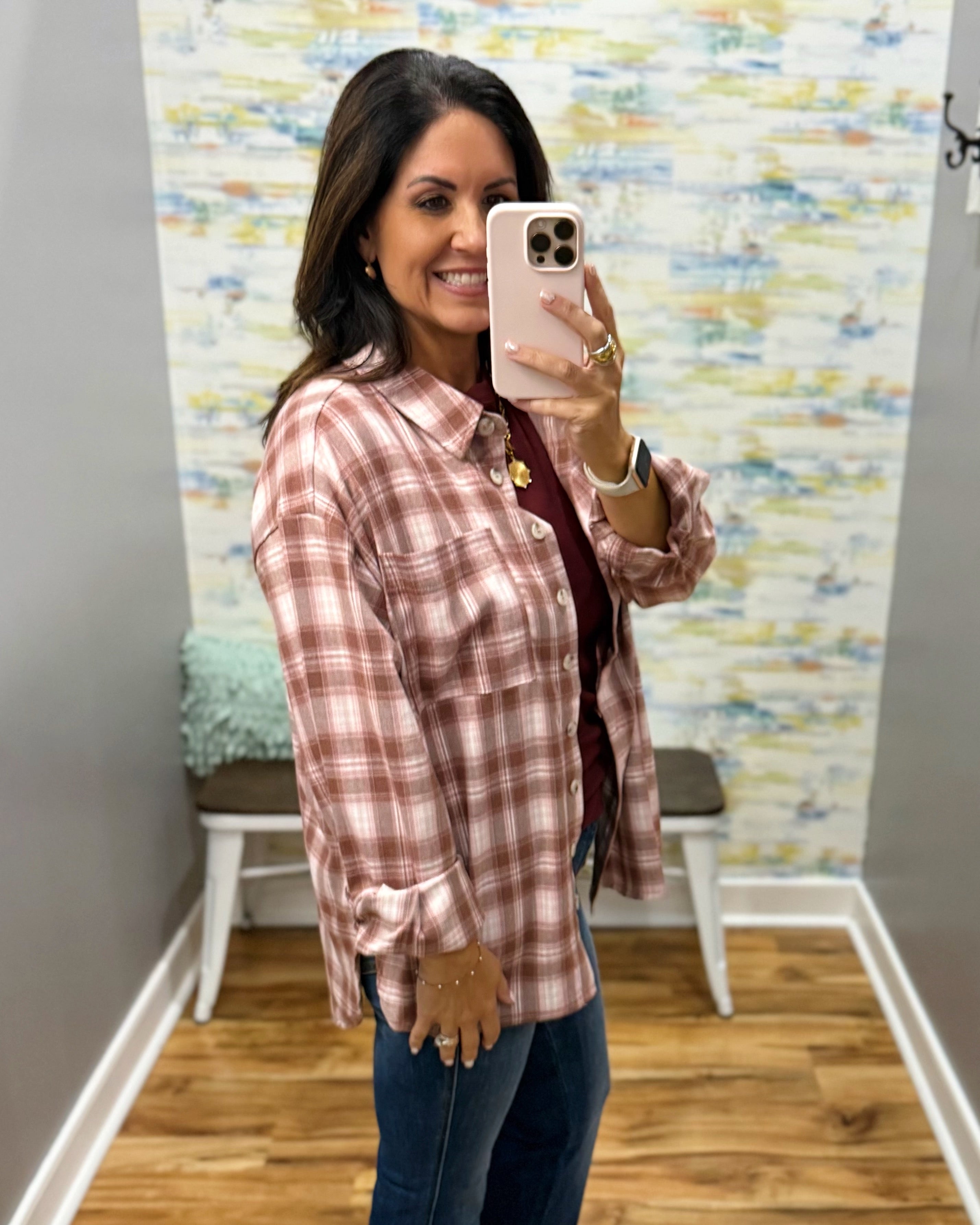A woman takes a mirror selfie wearing a dusty rose and white plaid button-down shirt over a dark red top and blue jeans, standing in a room with a patterned wallpaper background.