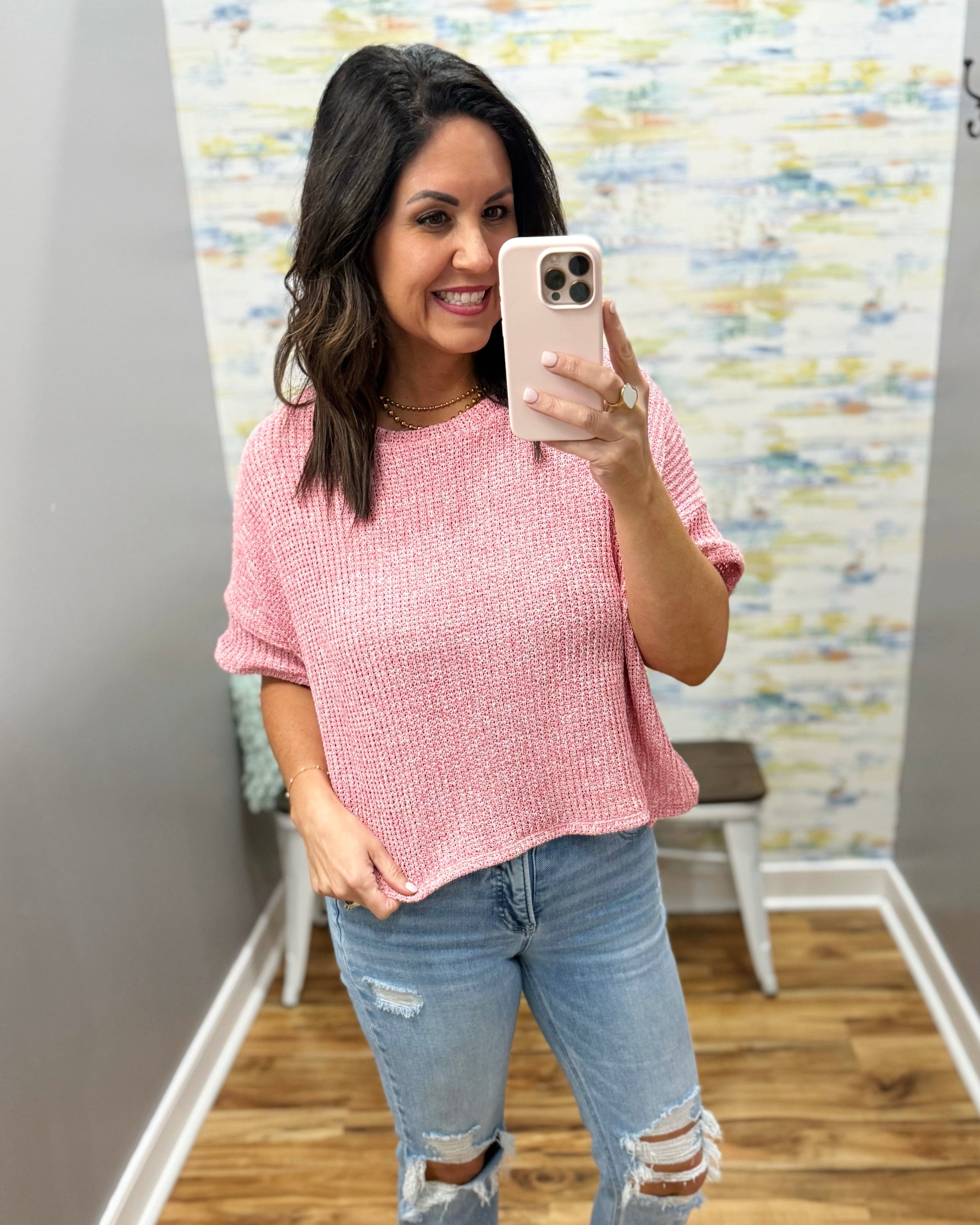 Woman taking a mirror selfie wearing a pink top and ripped jeans in a room with a colorful wall.