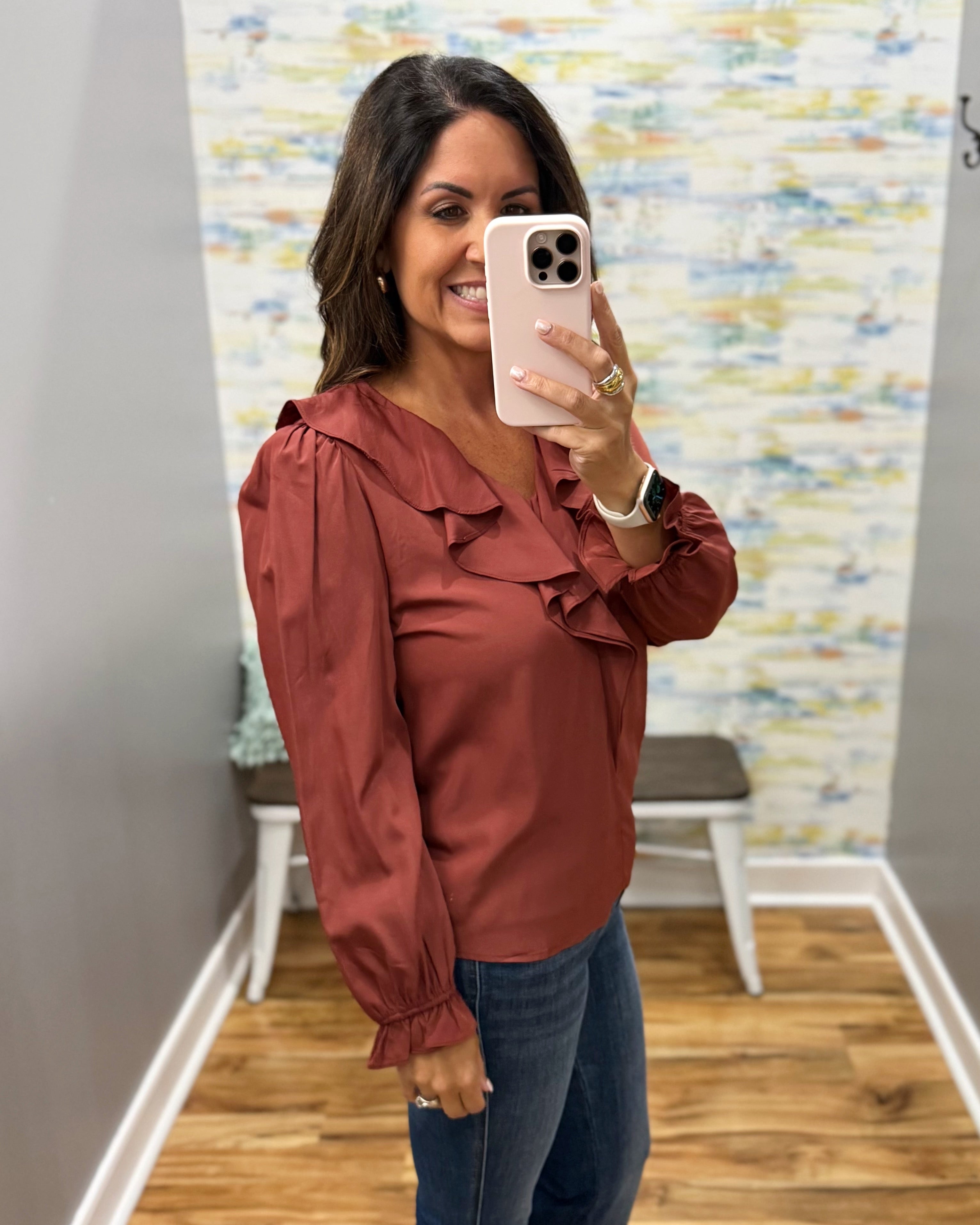 A woman takes a mirror selfie wearing a rust-colored long-sleeve blouse with ruffle details and blue jeans in a fitting room.