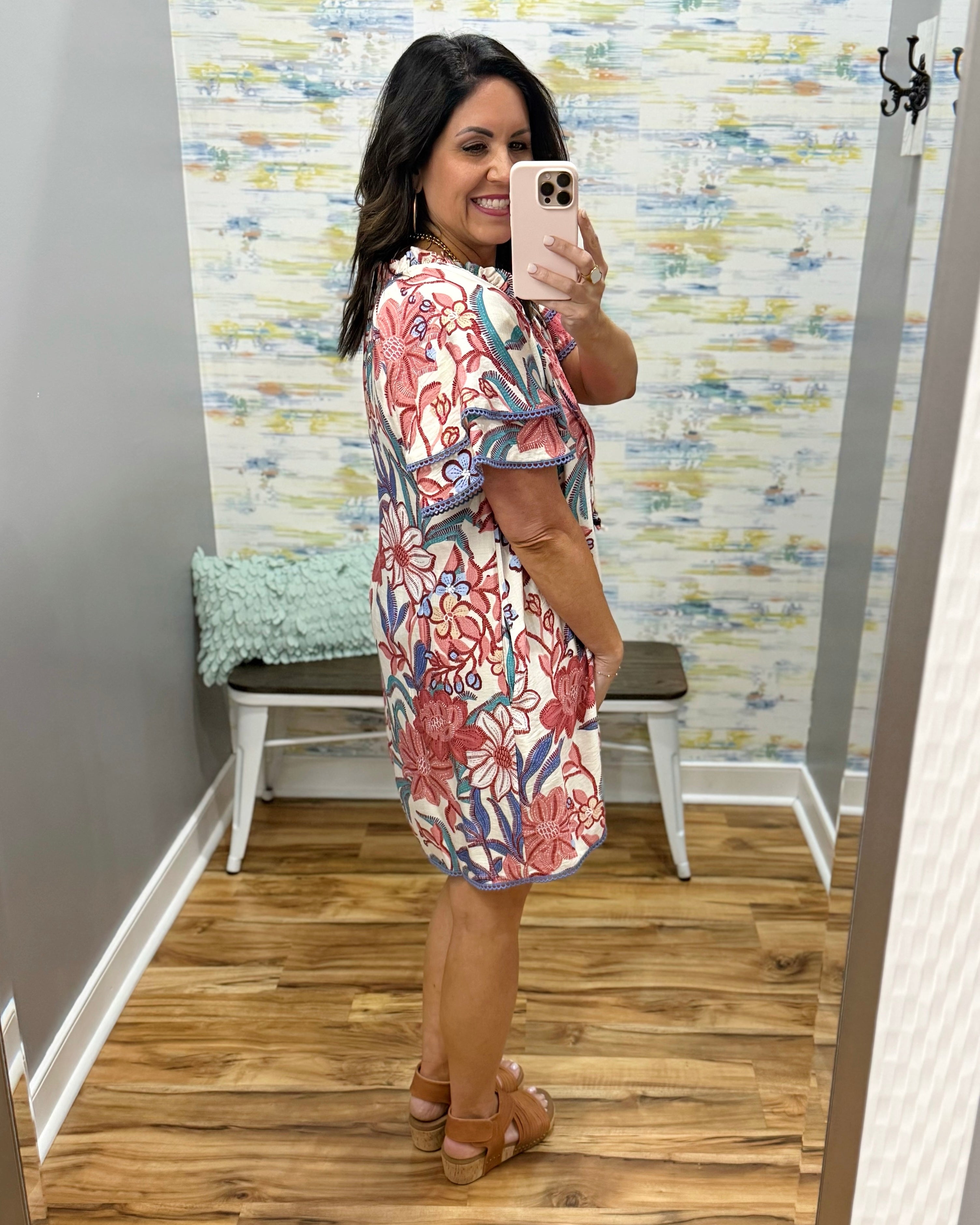 Woman taking a mirror selfie wearing a floral dress in a room with a patterned wall.
