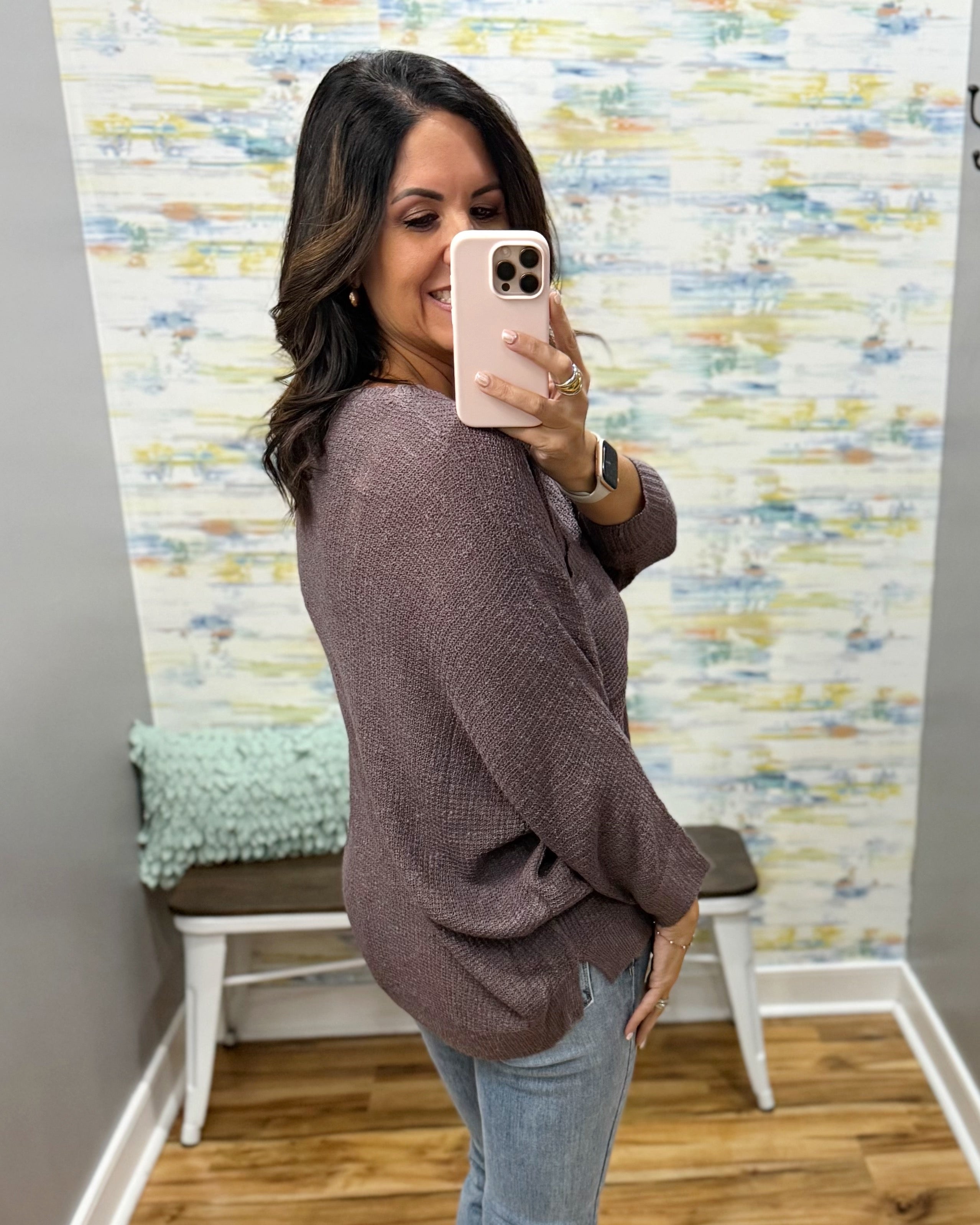 A woman takes a mirror selfie wearing a textured, waffle-knit long-sleeve sweater in a muted mauve-brown color, paired with light-wash blue jeans. The setting is a fitting room with a patterned wallpaper background and a bench with a mint green pillow.