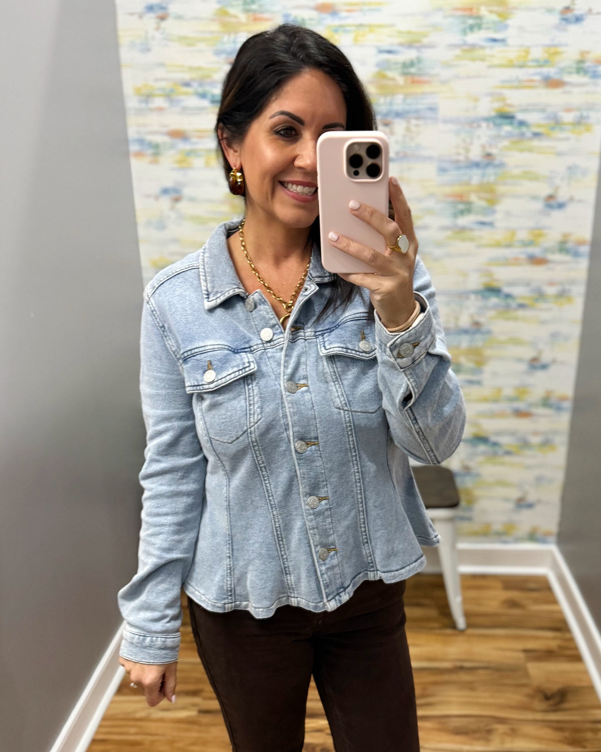 A woman taking a mirror selfie in a boutique wearing a light-wash blue denim jacket and dark brown pants.