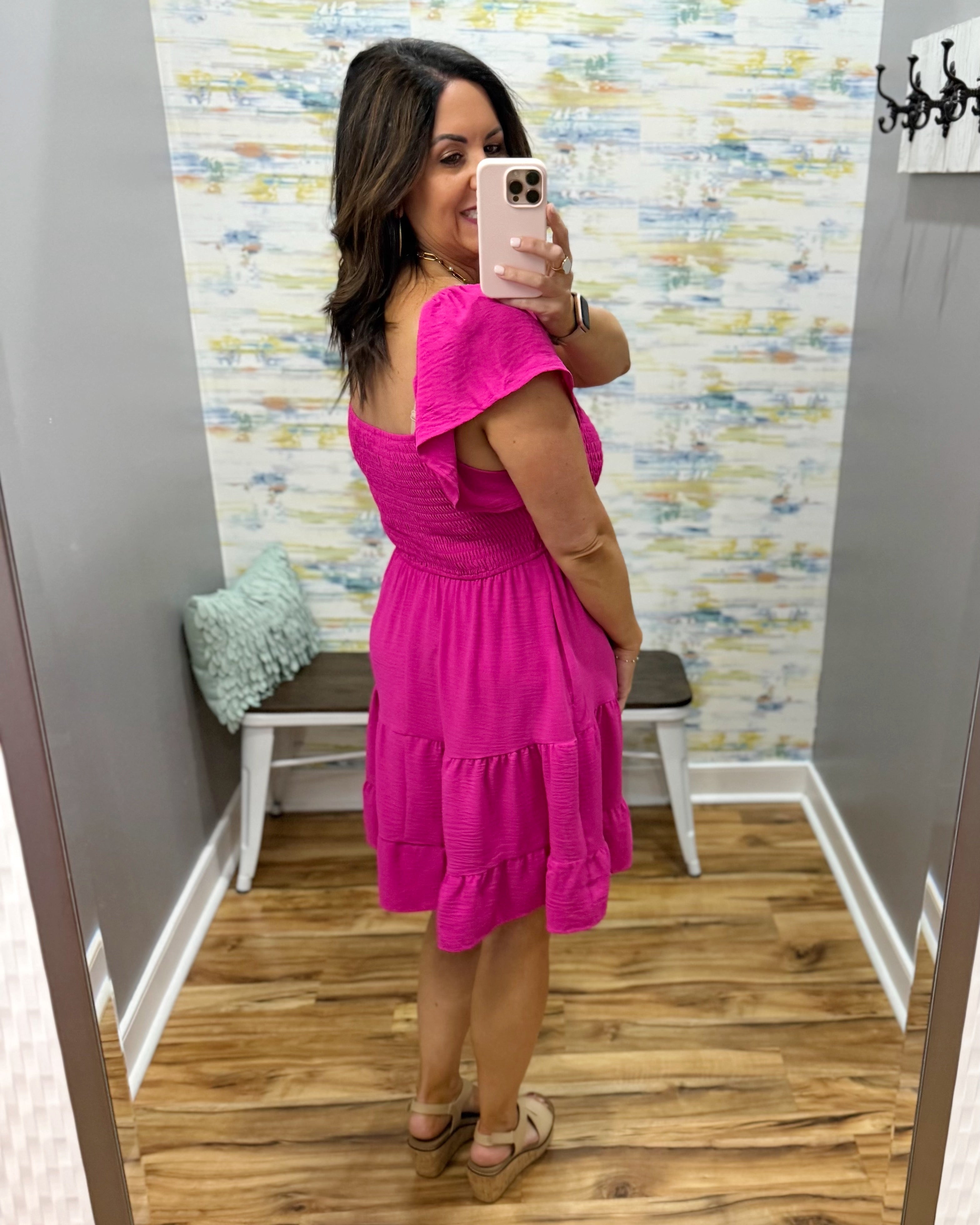 Woman taking a mirror selfie wearing a bright pink dress in a room with colorful wallpaper.