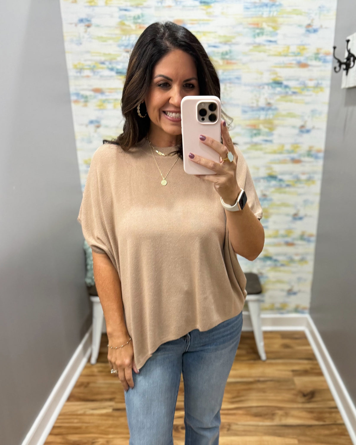 A woman takes a mirror selfie wearing a beige short-sleeve knit top and blue jeans in a fitting room with a patterned wallpaper background.