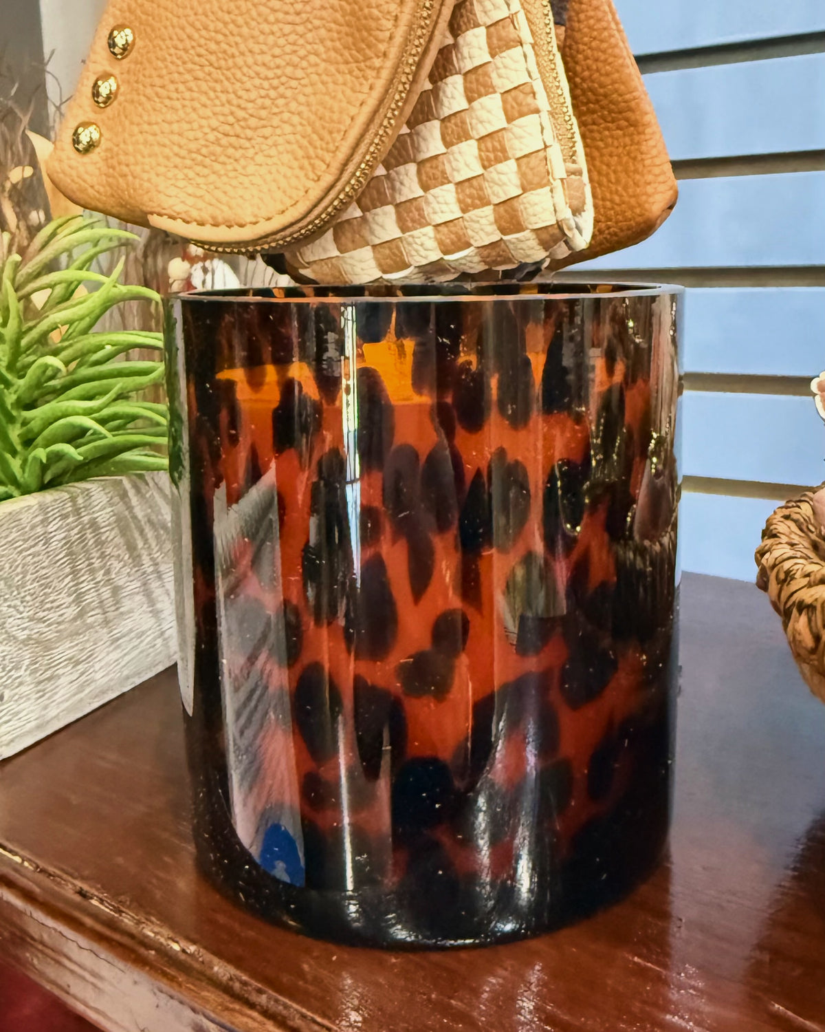 A glossy cylindrical glass sweet grace #64 candle with a black, brown, and orange leopard print pattern, styled on a wooden table with a handbag draped over the top.