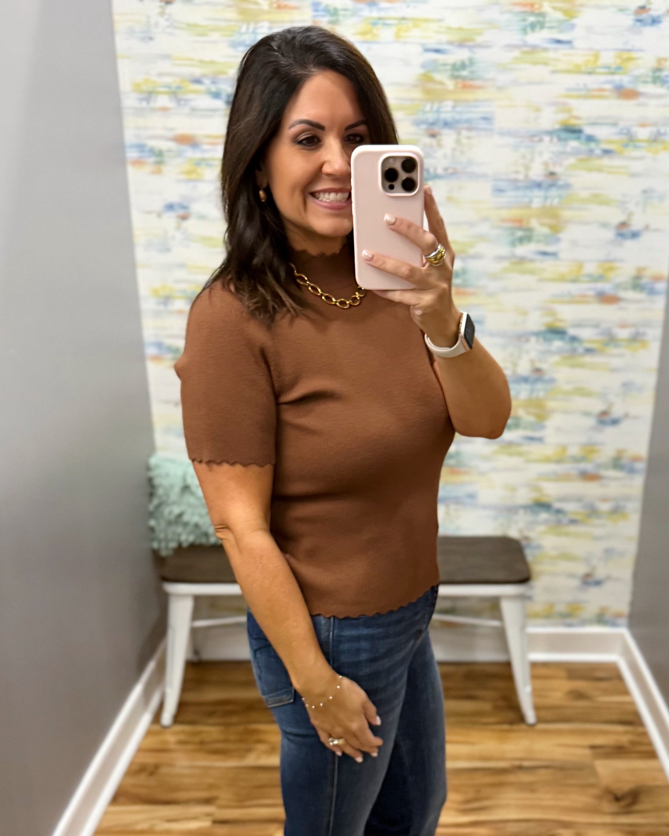 A woman takes a mirror selfie in a fitting room wearing a brown short-sleeve knit top with scalloped edges and blue jeans.