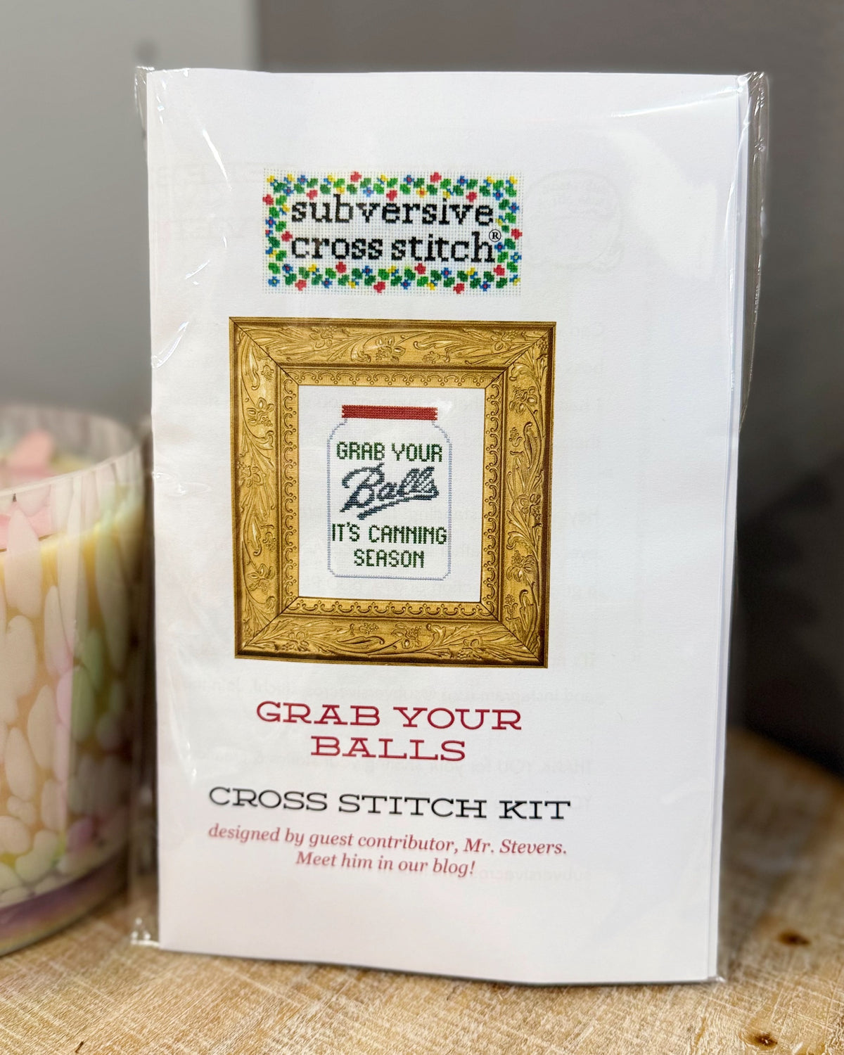 A packaged cross stitch kit labeled 'subversive cross stitch' with the design text 'Grab Your Balls / It's Canning Season,' shown in clear plastic wrapping on a tabletop next to a candle.