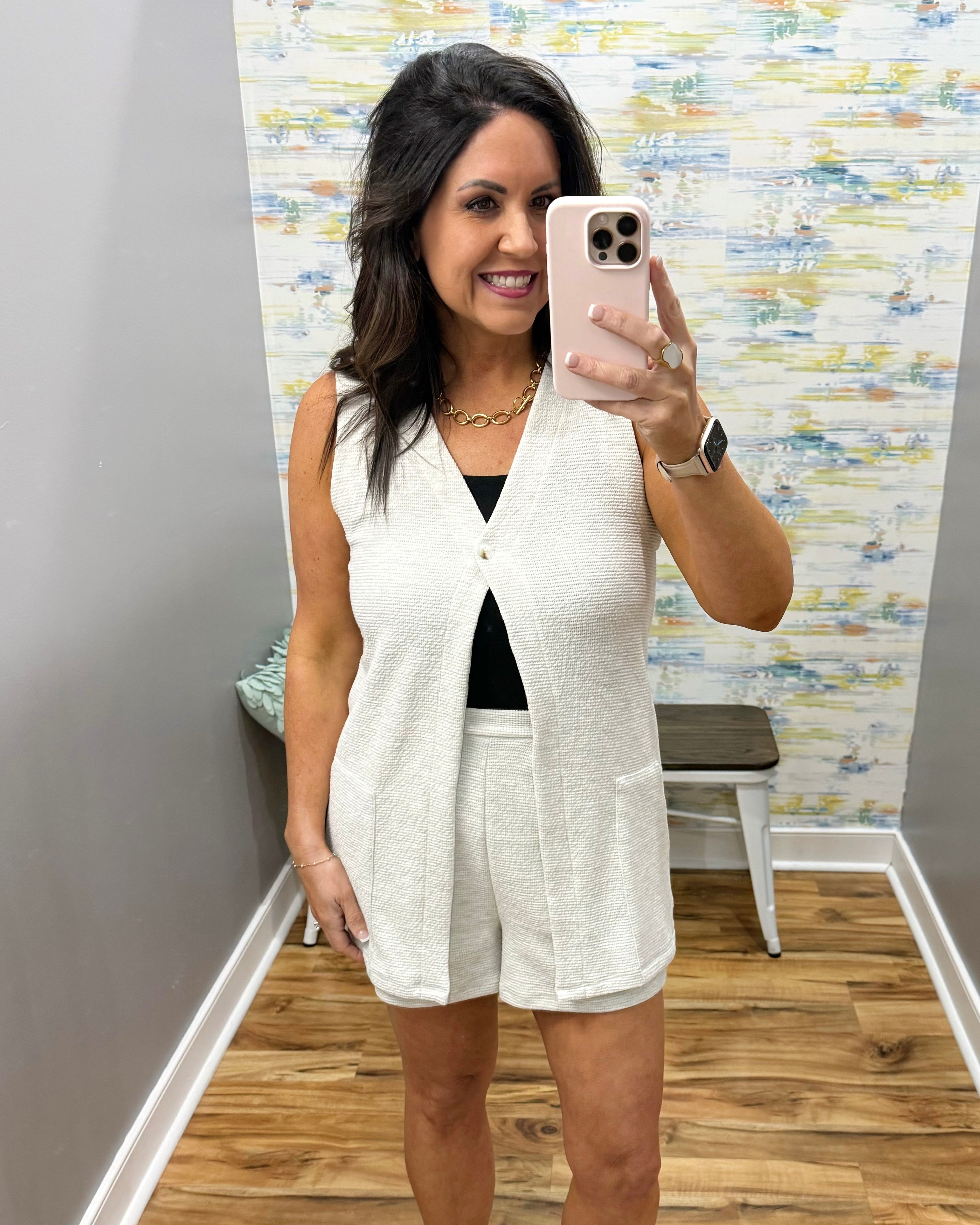 Woman taking a mirror selfie wearing a white vest and shorts in a room with patterned wallpaper.