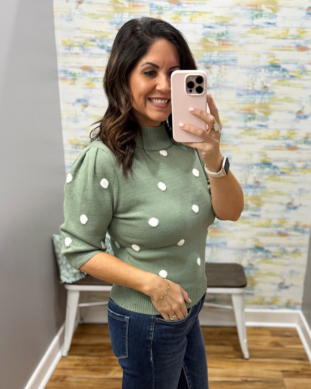 A woman takes a mirror selfie wearing a sage green mock-neck knit sweater with white polka dots and dark blue jeans, standing in front of a patterned wallpaper background.