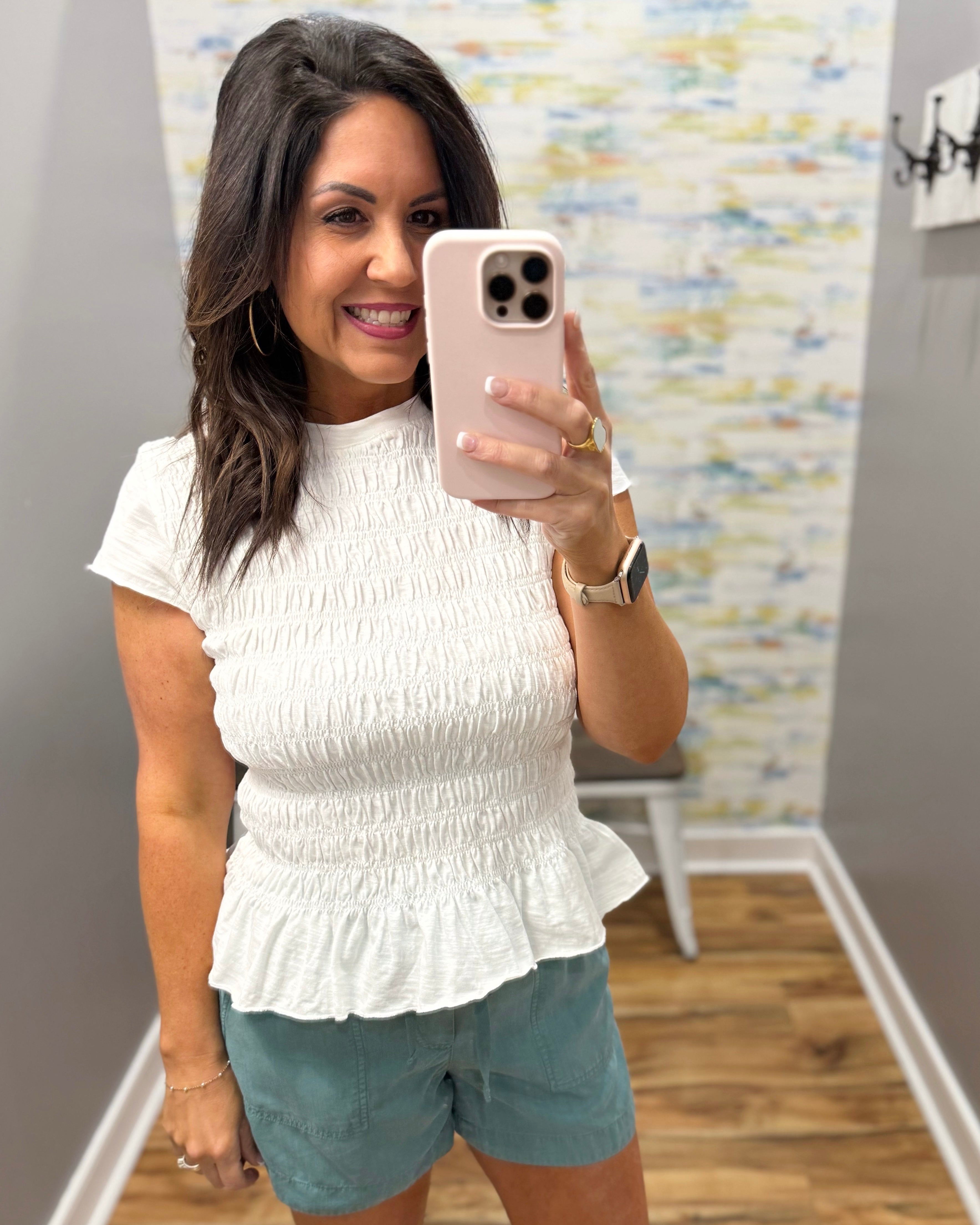 Woman taking a mirror selfie wearing a white top and light blue shorts in a room with a colorful wall.