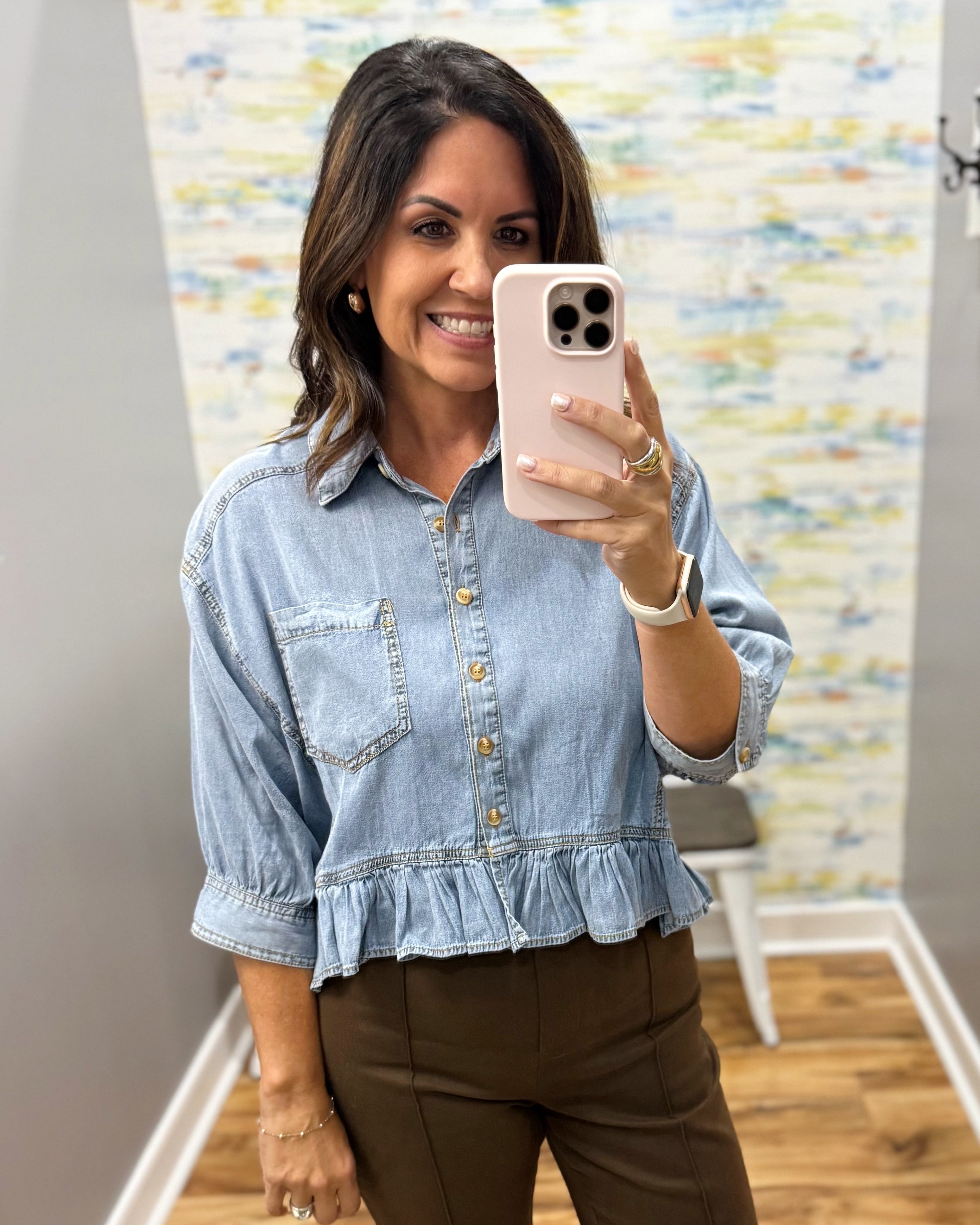 A woman taking a mirror selfie wearing a light-wash blue denim shirt with rolled sleeves, gold-tone buttons, and a ruffled hem, paired with brown pants.