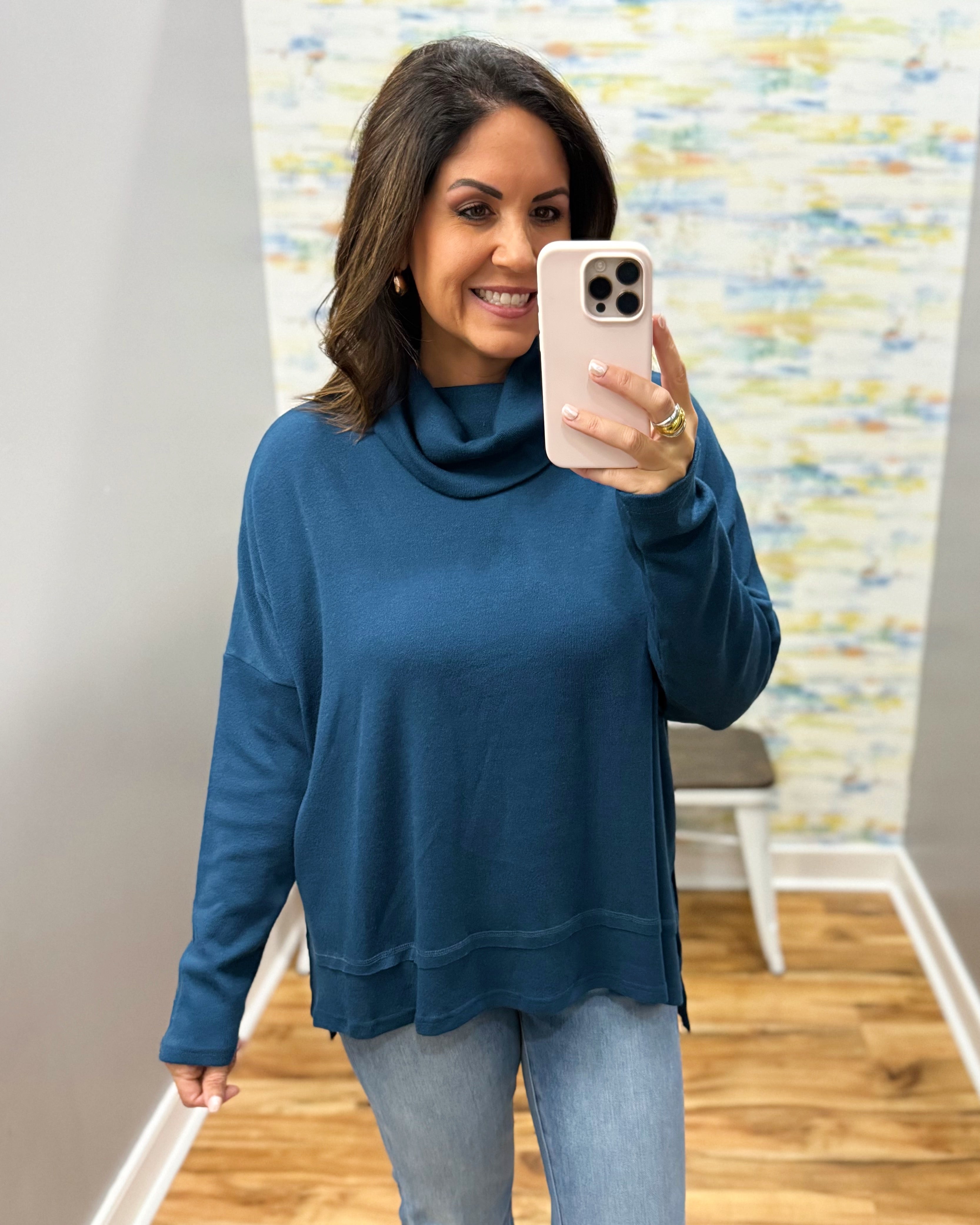 A woman takes a mirror selfie wearing a slate blue cowl-neck long-sleeve top and light-wash jeans in a home setting with a wood floor and abstract wall art.
