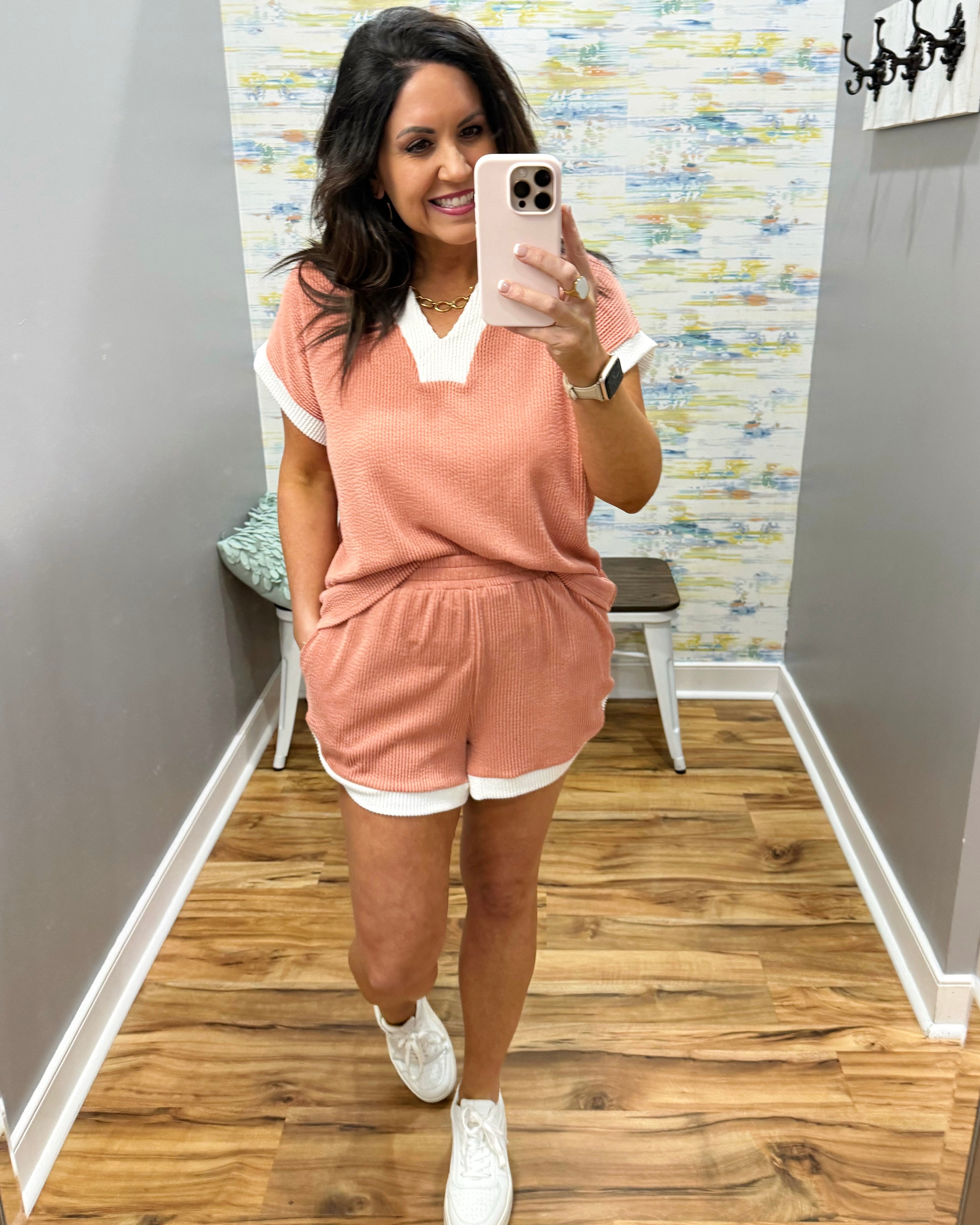 Woman taking a mirror selfie wearing a pink outfit in a room with wooden flooring and a colorful wall.
