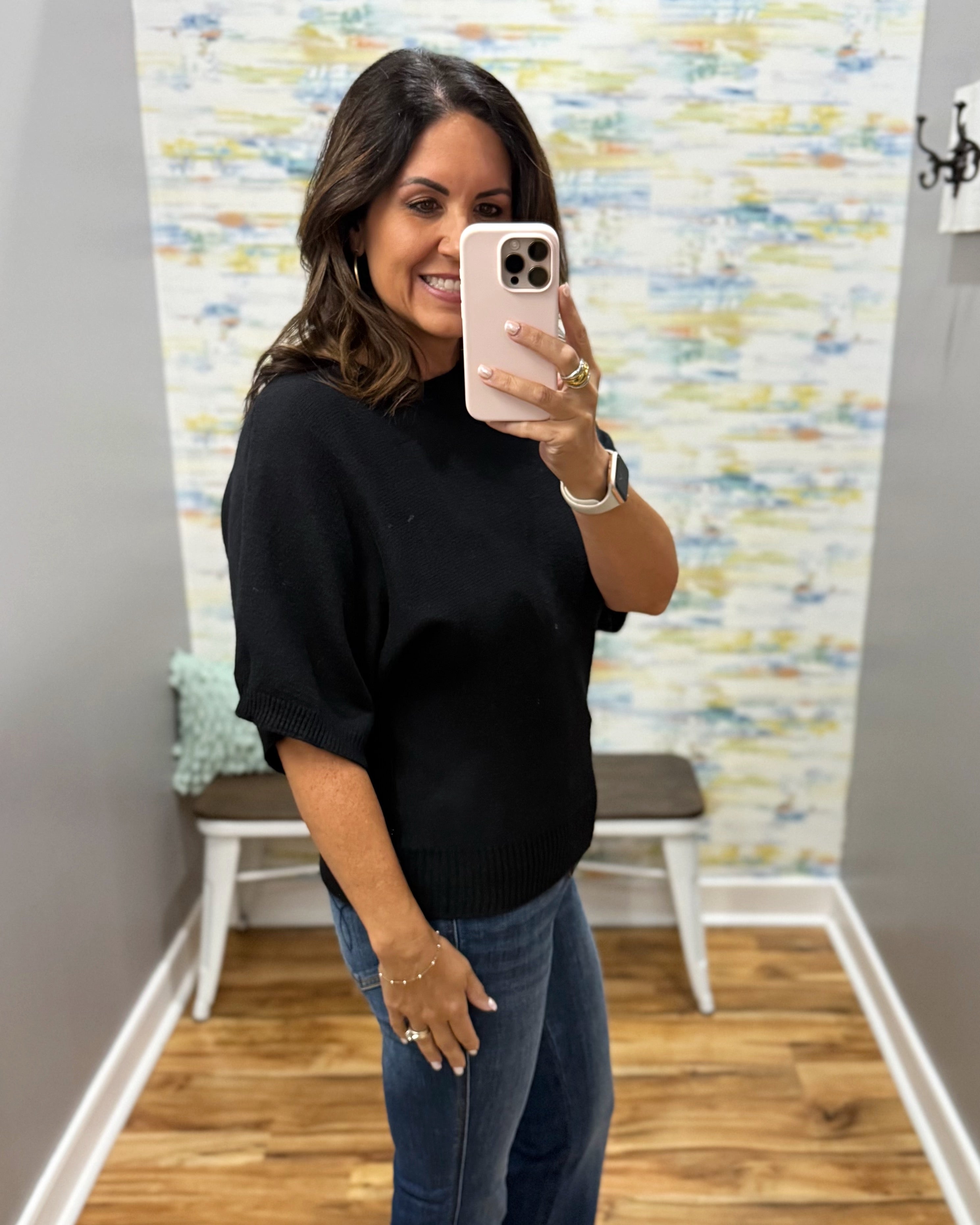 A woman takes a mirror selfie in a fitting room wearing a black short-sleeve knit sweater and blue jeans.