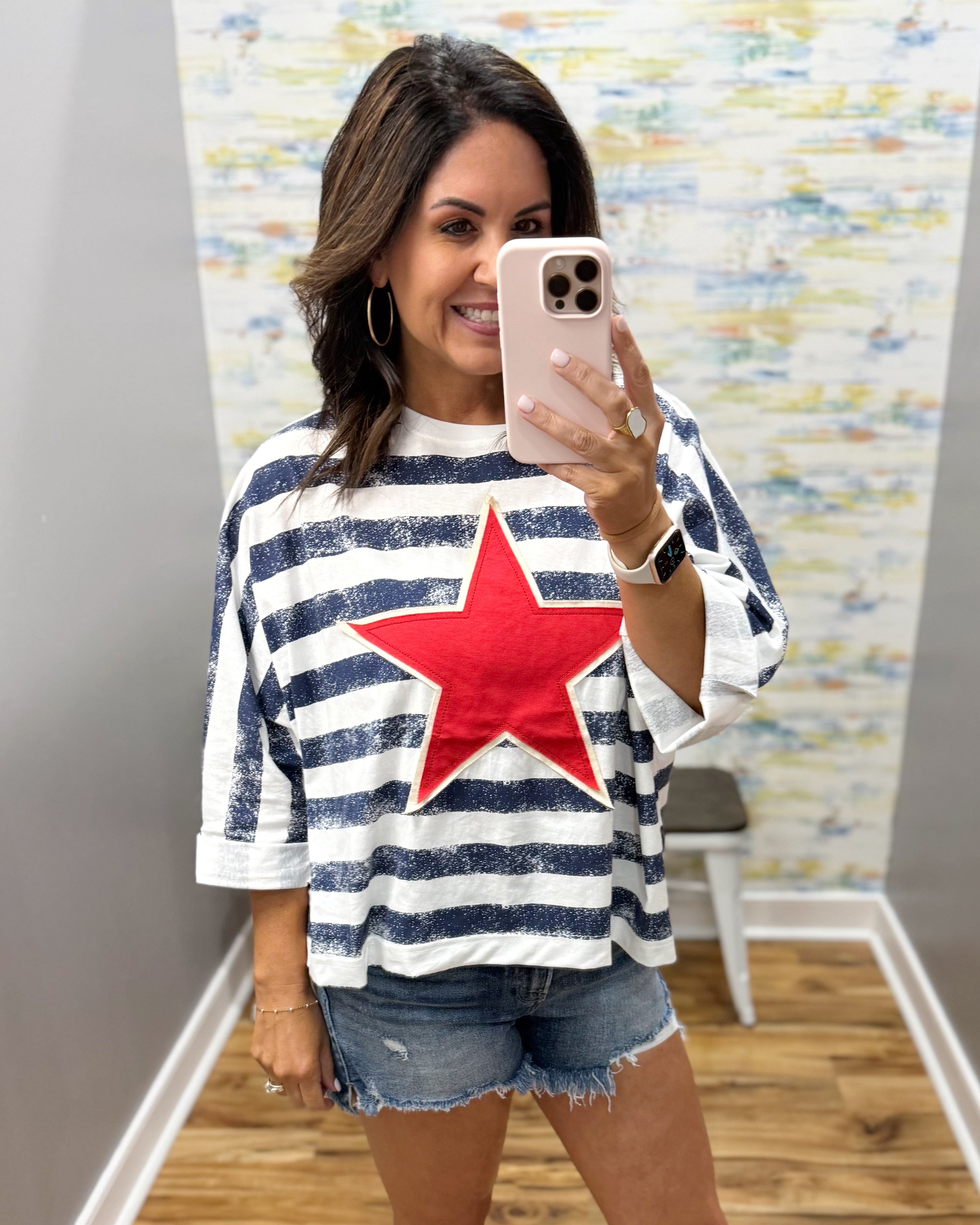 Woman wearing a striped shirt with a red star, taking a mirror selfie.