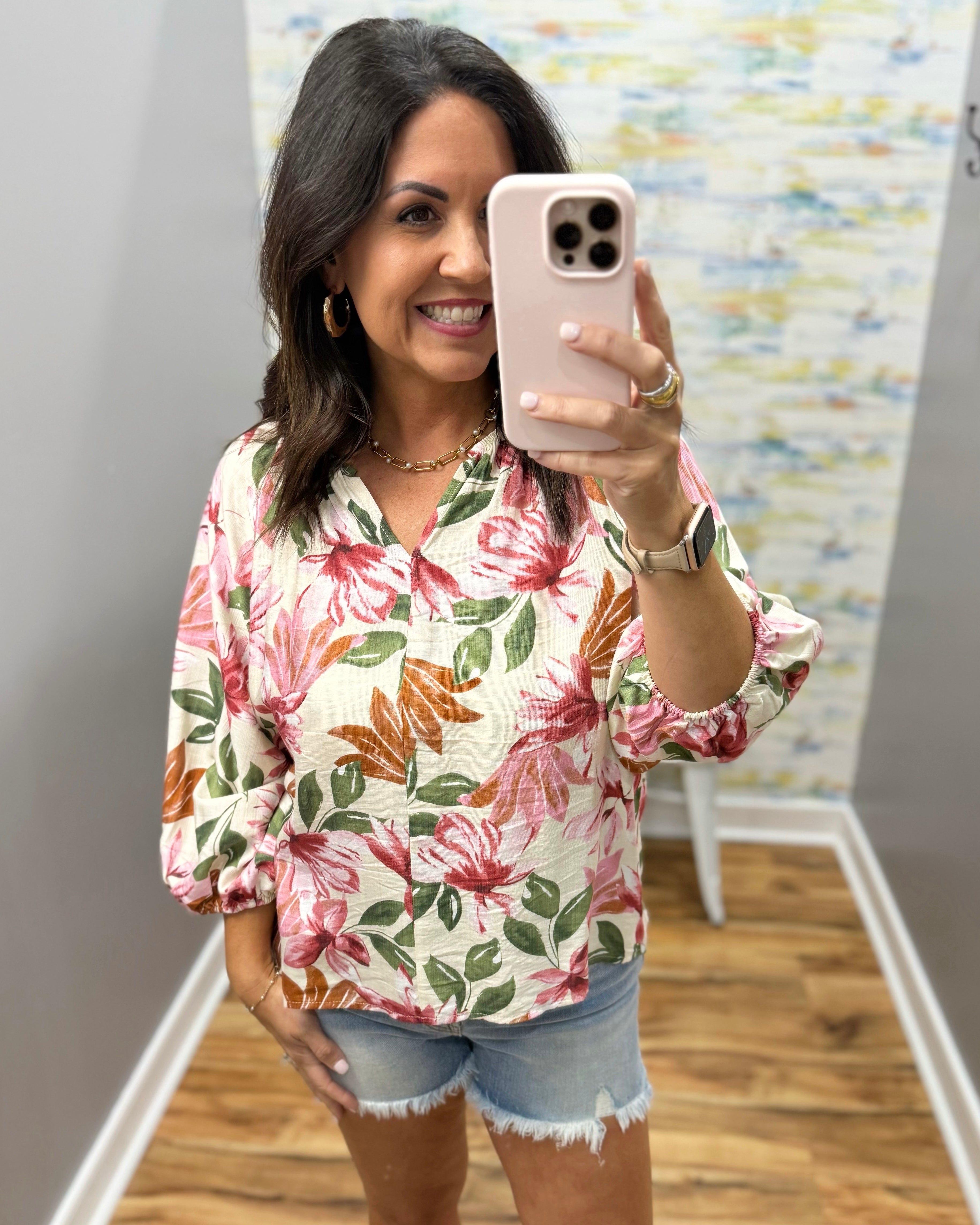 Woman taking a mirror selfie wearing a floral blouse and denim shorts.