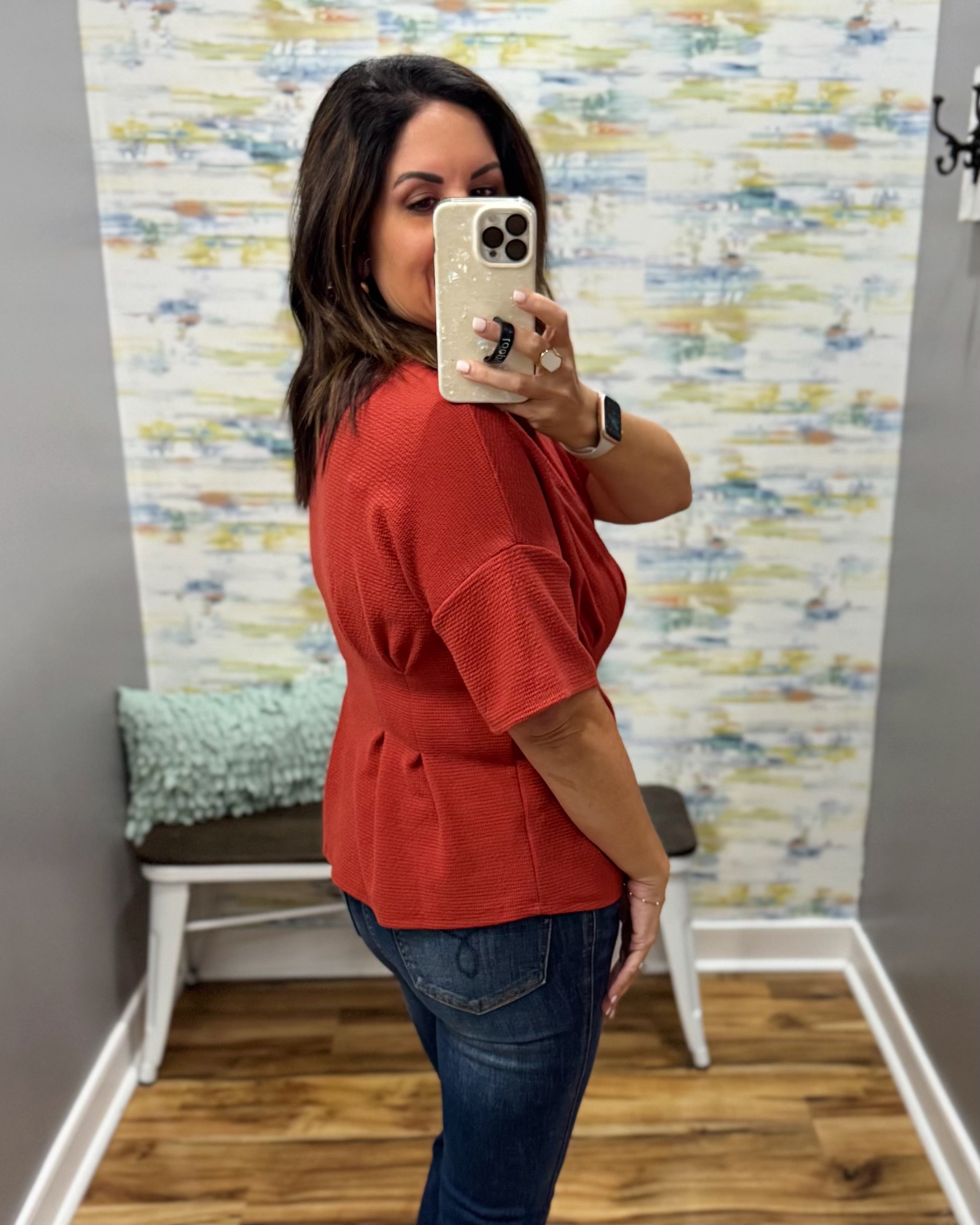 A woman takes a mirror selfie in a fitting room wearing a rust-orange short-sleeve peplum top and dark blue jeans.