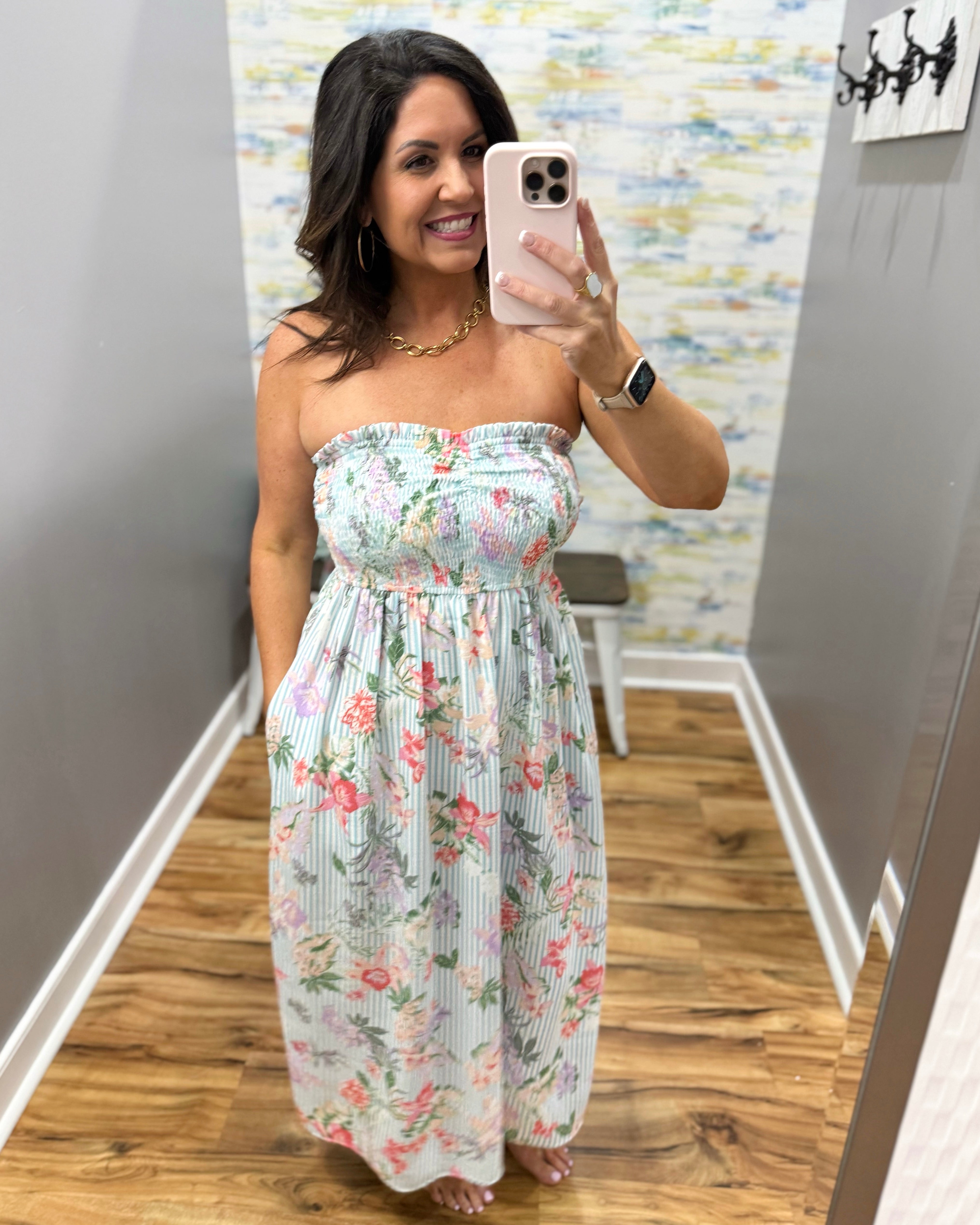 Woman taking a mirror selfie wearing a floral strapless dress in a room with patterned wallpaper.