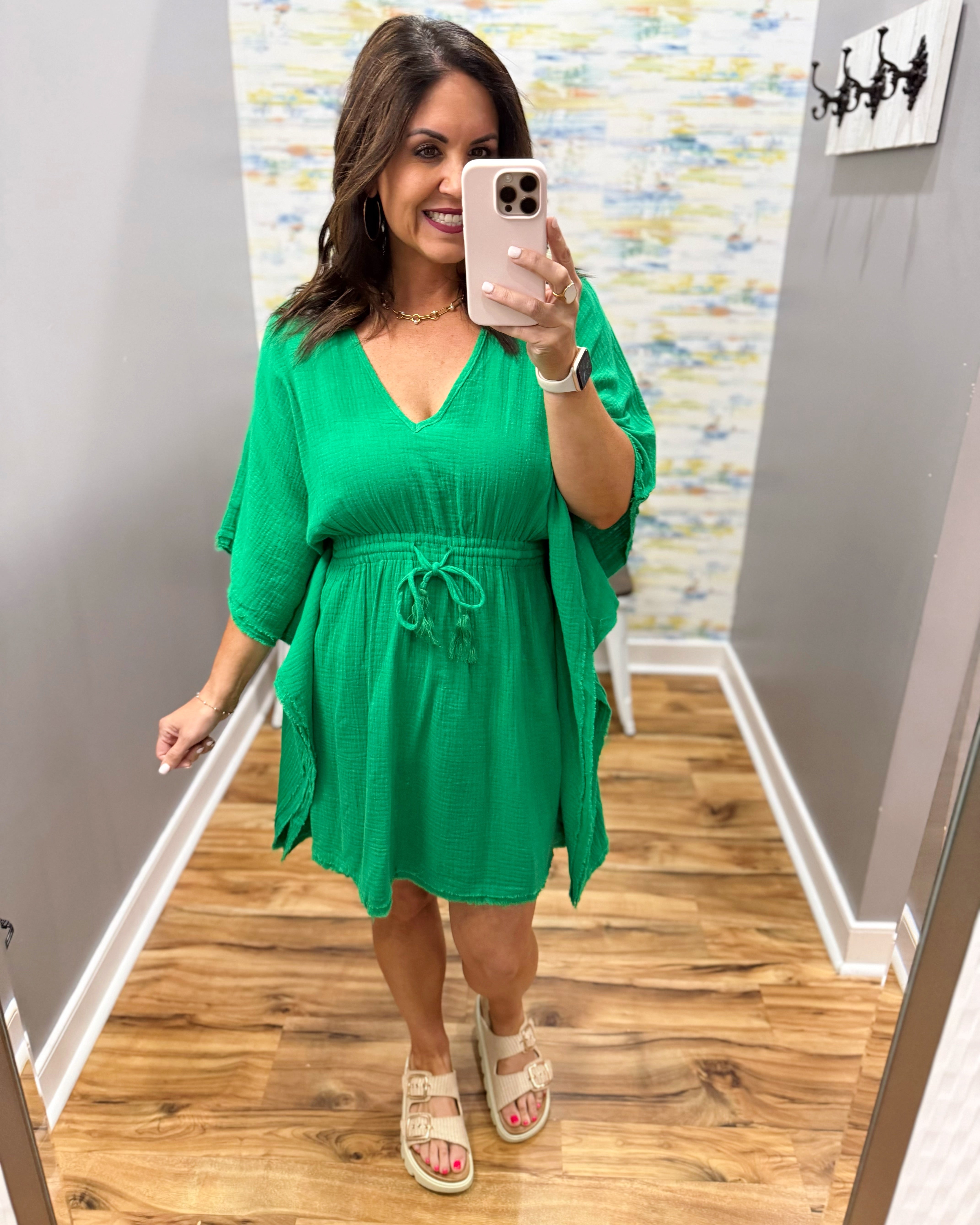 Woman wearing a green dress taking a mirror selfie in a room with wooden flooring and a colorful wall.