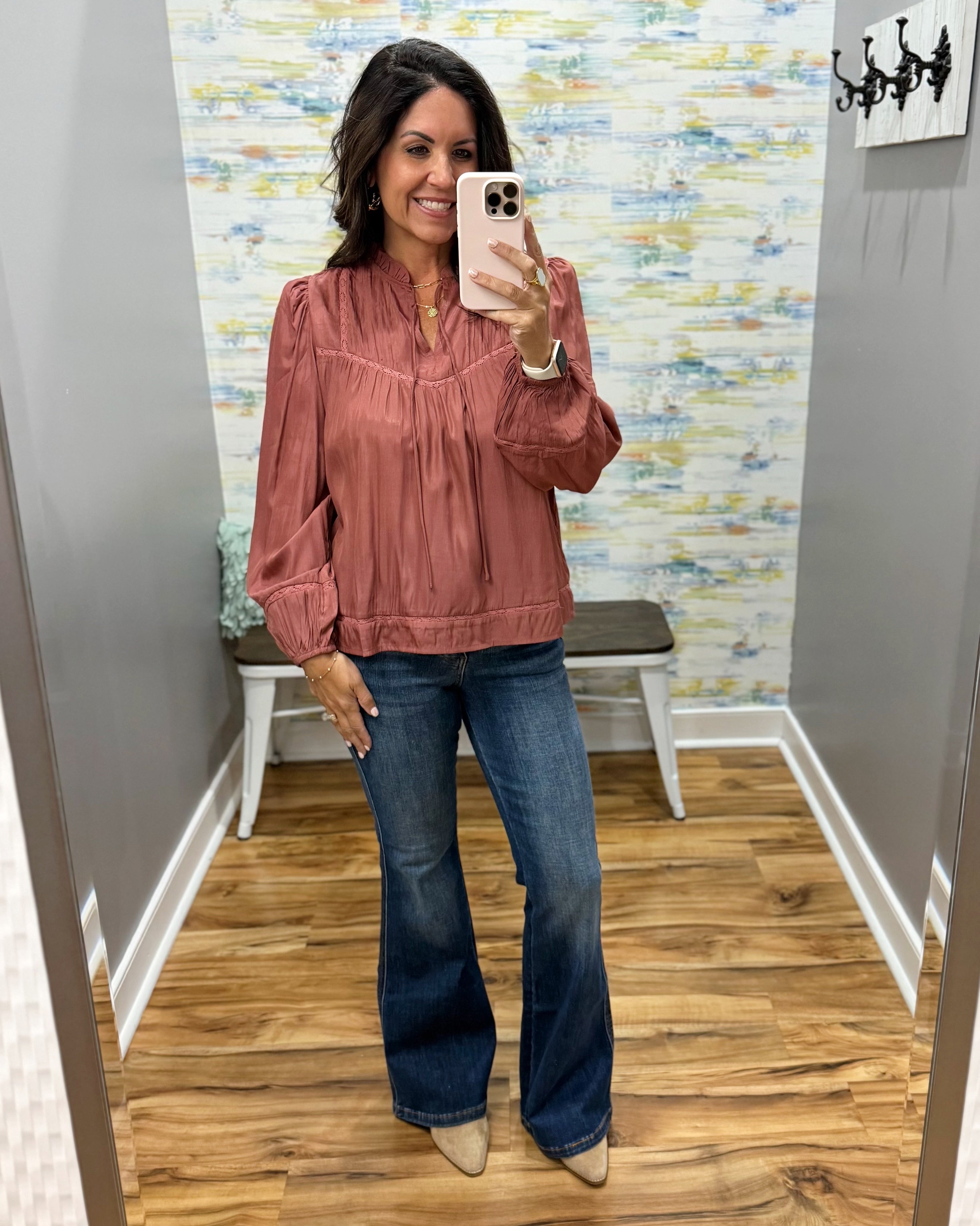 A woman takes a mirror selfie wearing a dusty rose long-sleeve blouse with a smocked chest and blue flare jeans in a fitting room.