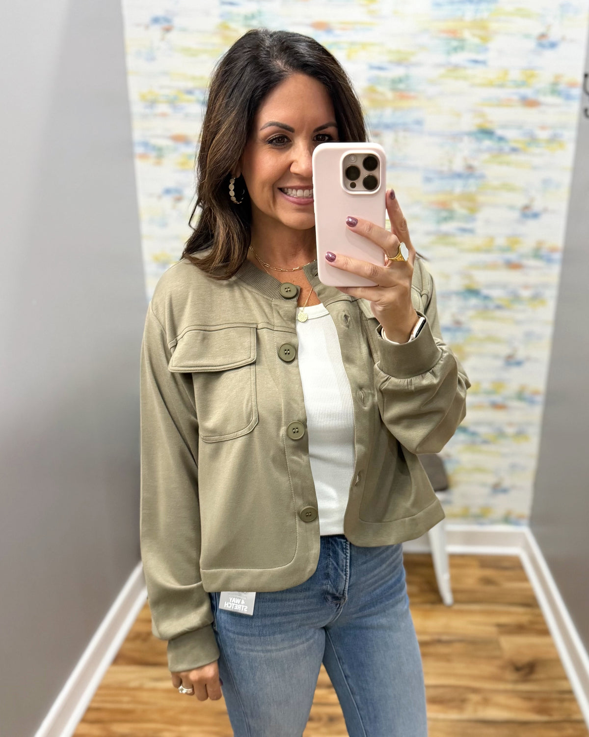 A woman takes a mirror selfie wearing a cropped sage green utility jacket over a white top and blue jeans.