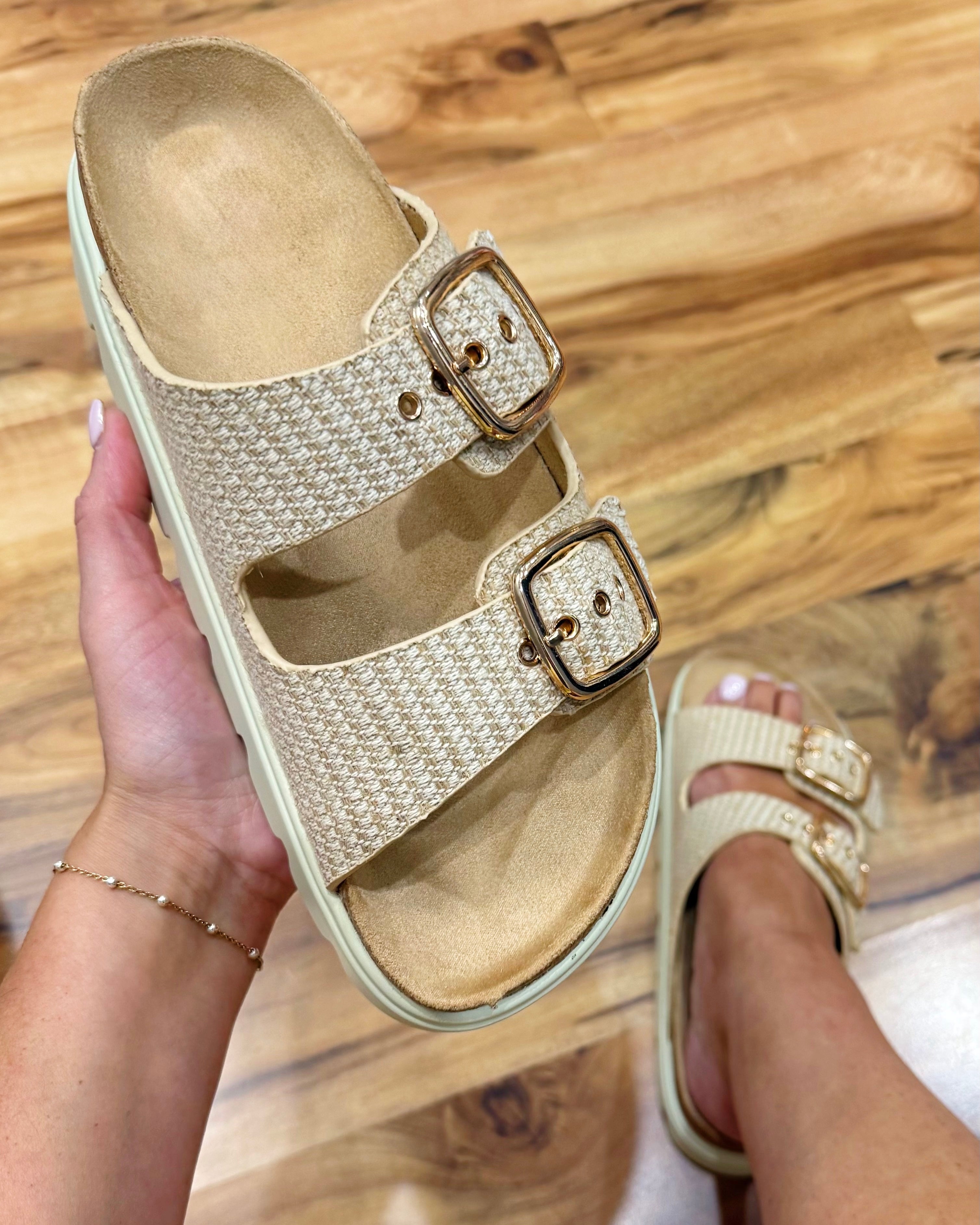 Beige sandals with gold buckles held by a hand on a wooden floor.