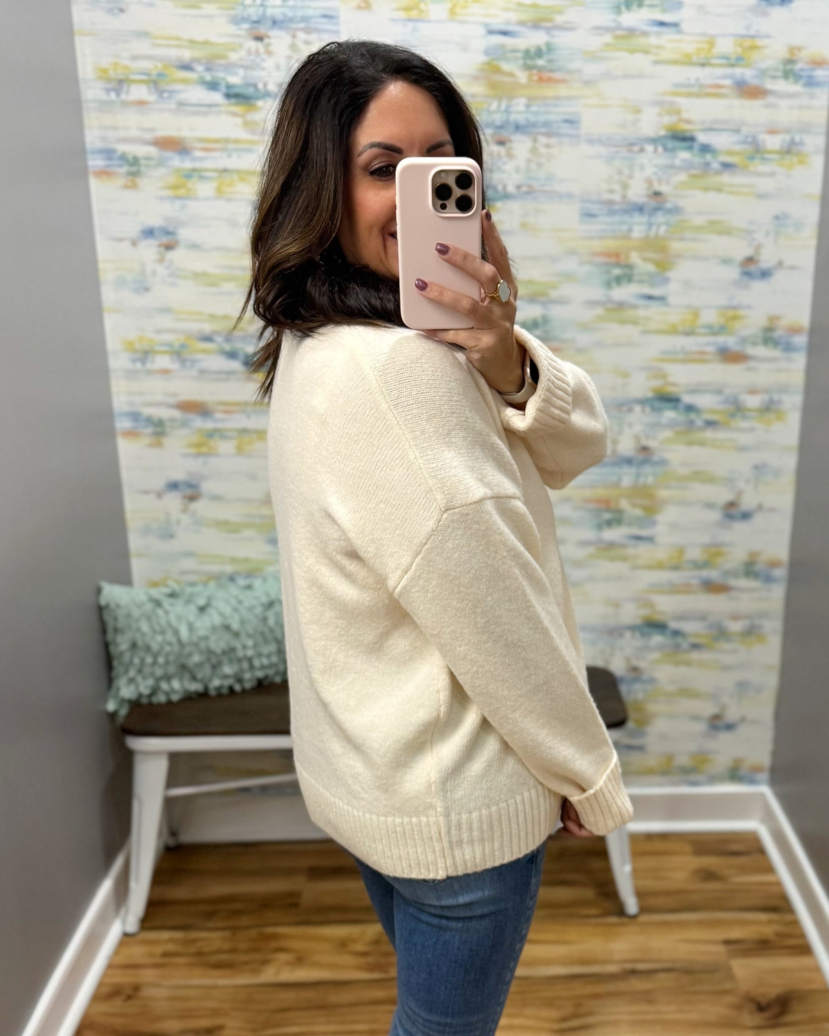 A woman takes a mirror selfie wearing a cream-colored knit sweater and blue jeans in front of a patterned wallpapered wall.