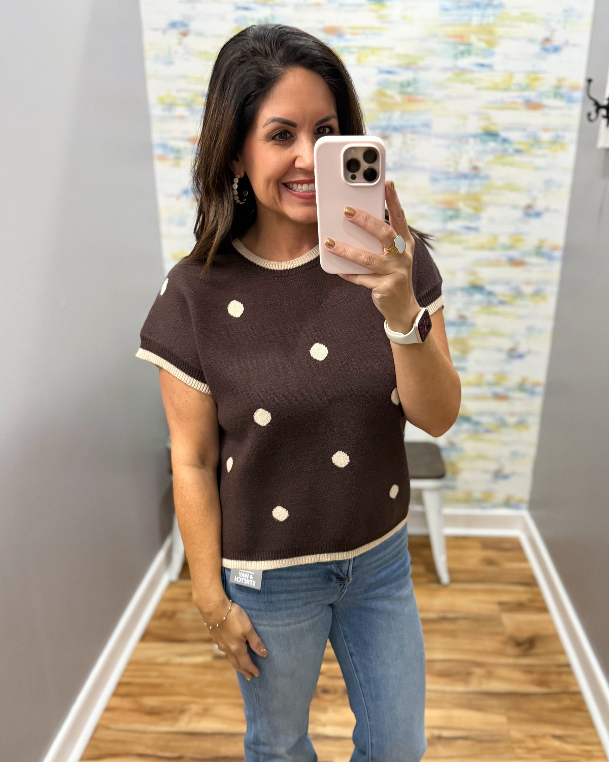 A woman takes a mirror selfie wearing a brown short-sleeve knit top with cream polka dots and trim, paired with blue jeans in an indoor room with wood floors and a patterned wall.