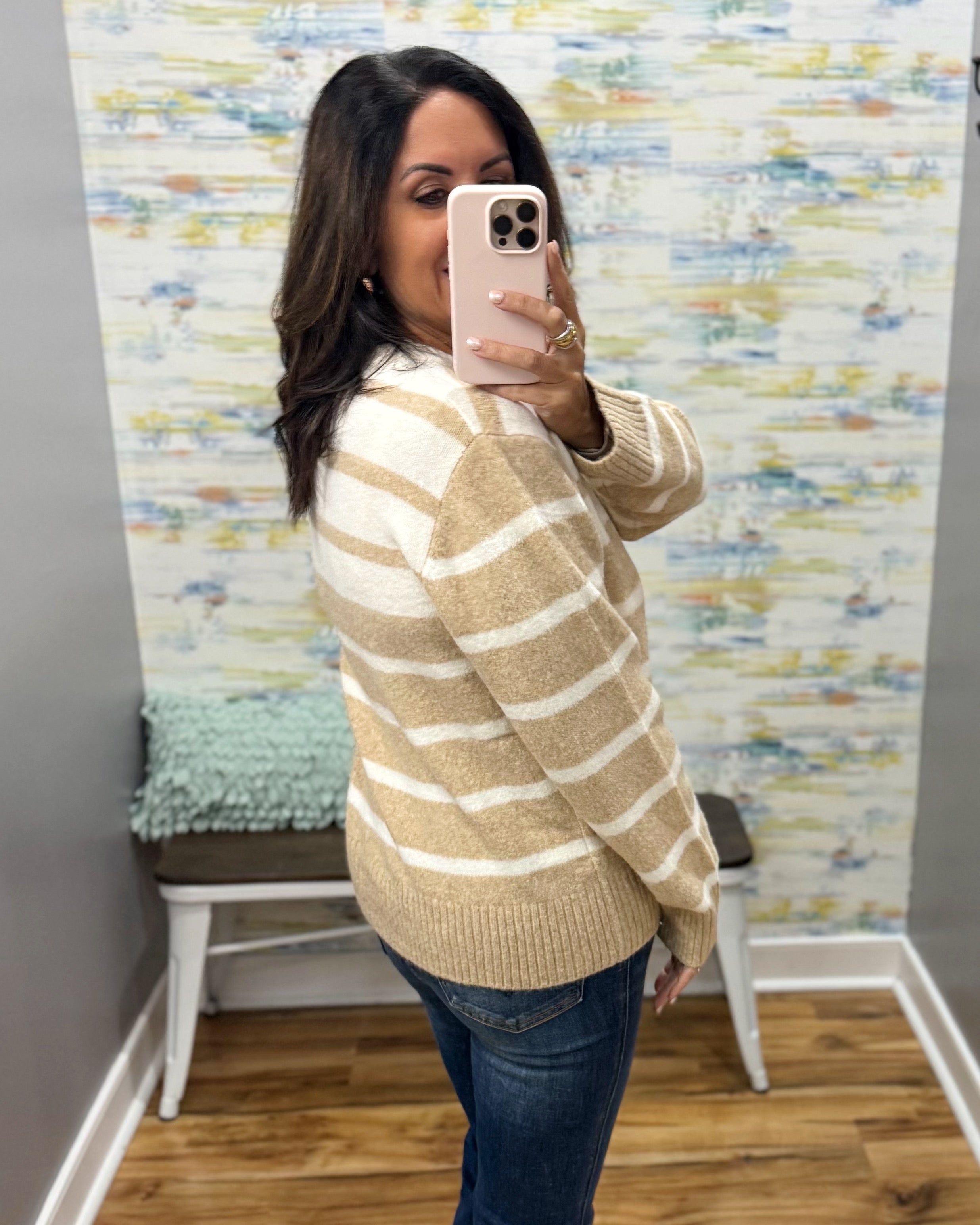 A woman takes a mirror selfie in a fitting room wearing a beige and white striped knit sweater and blue jeans.